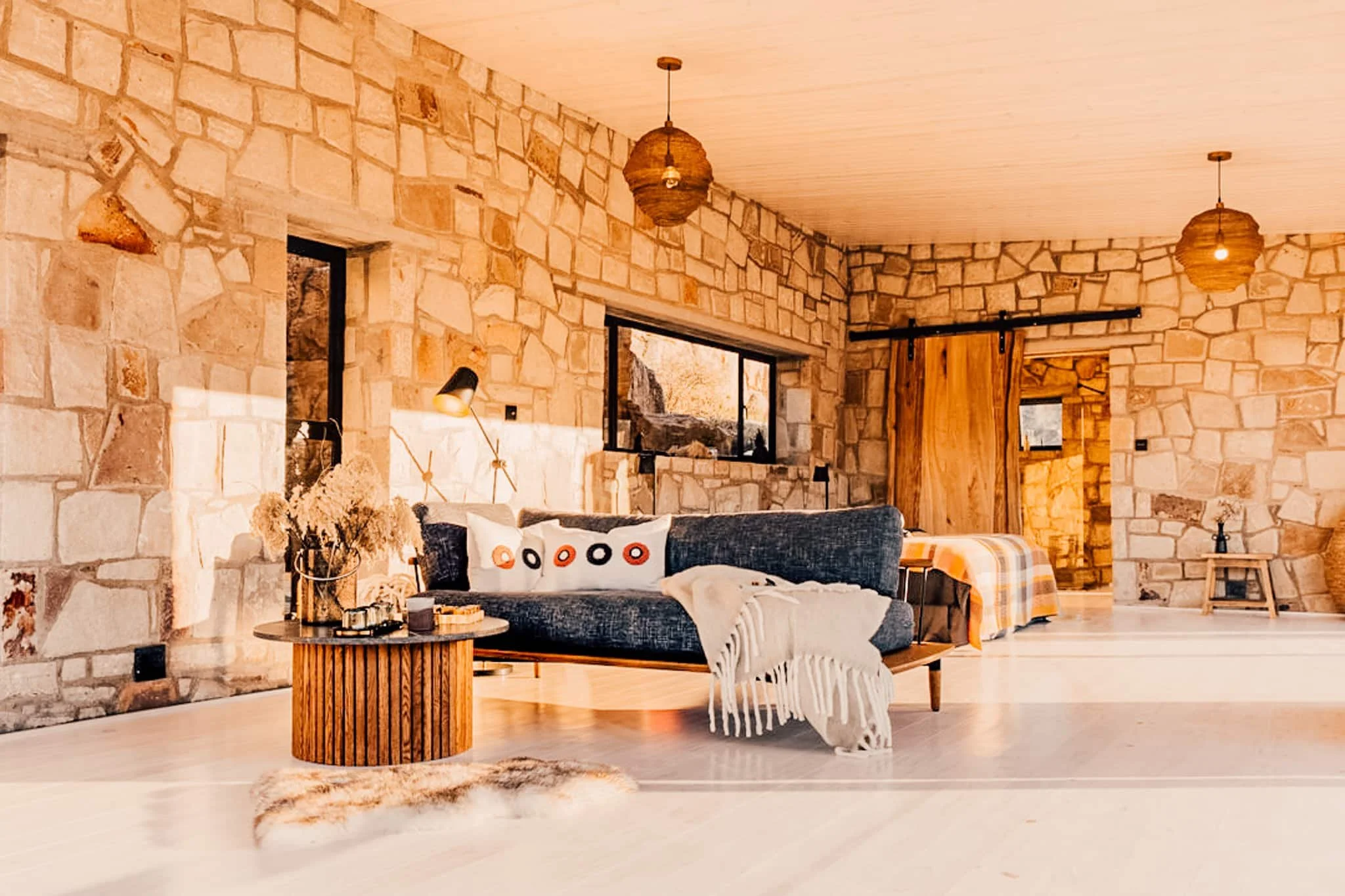 Cozy living room with beige stone walls, wooden ceiling, and modern decor. Features a dark gray sofa with decorative pillows, a round wooden coffee table, and a plush rug nearby. Hanging wicker pendant lights and large windows letting in natural light add to the warm atmosphere.