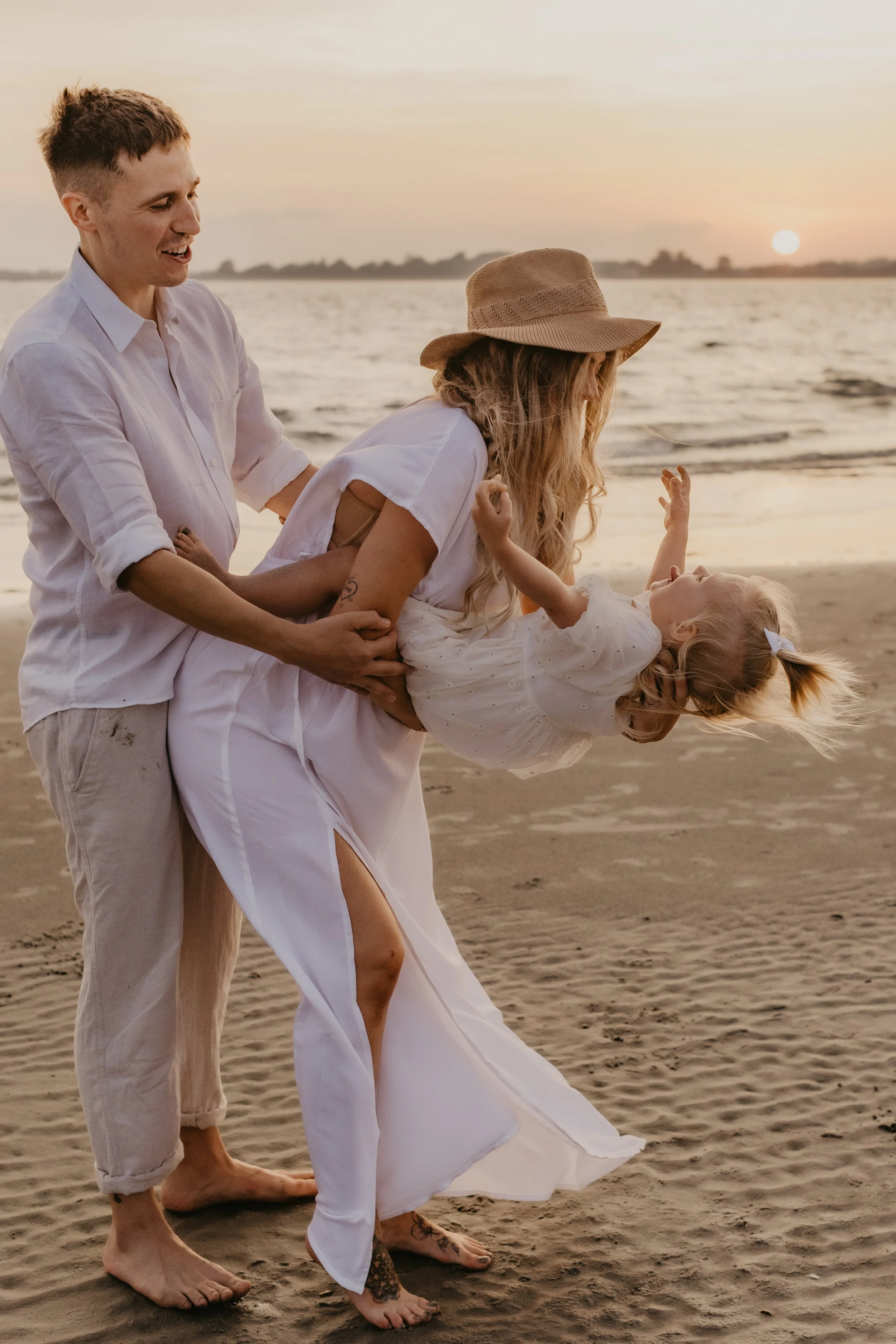 A family of three on the beach at sunset, with a man and woman playing with a young girl who is turned upside down and smiling.