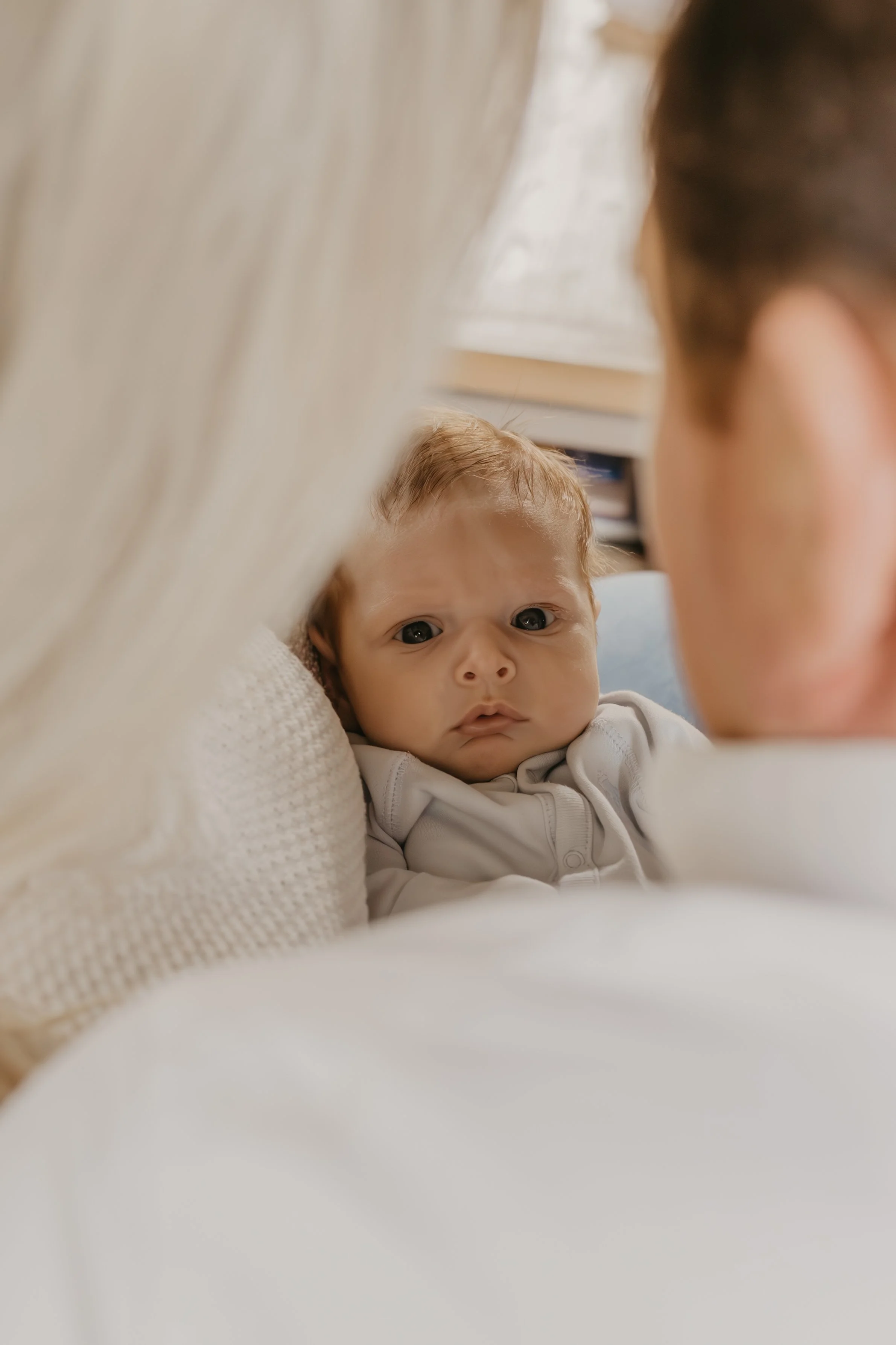 A close-up of a baby with light brown hair and big eyes, looking curiously at a woman with dark hair who is partially visible in the foreground, against a cozy indoor setting with neutral-colored furniture.