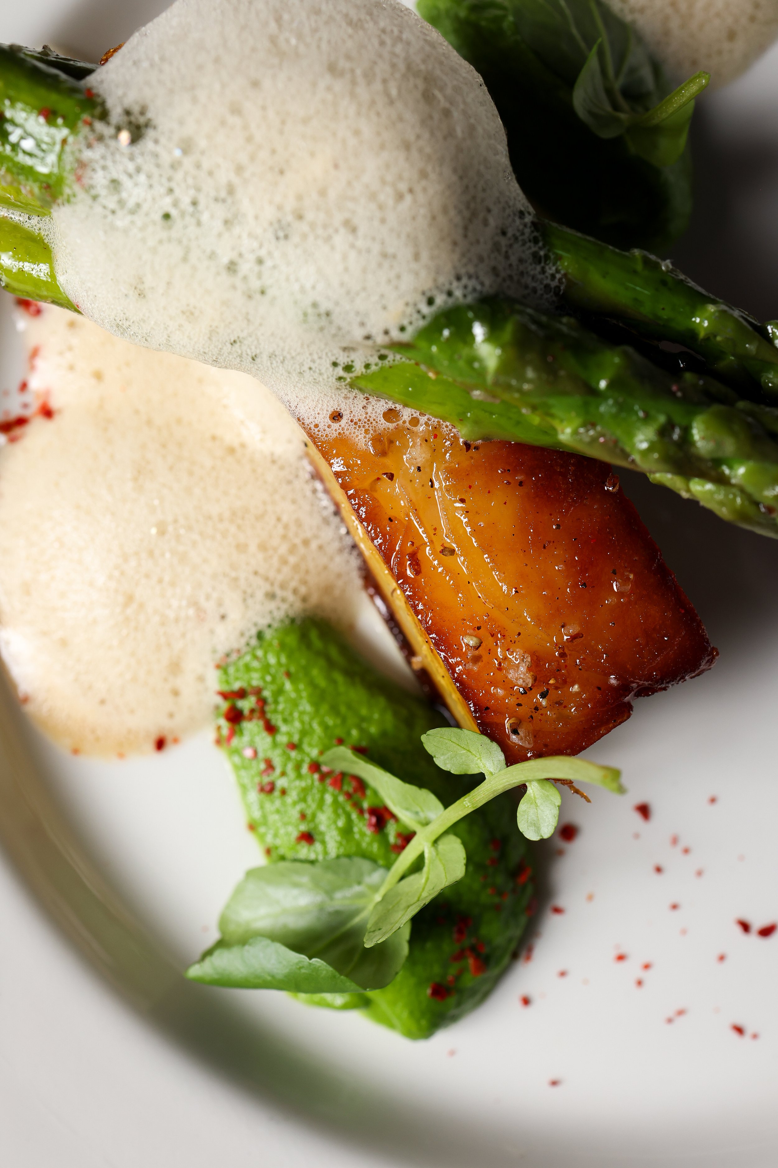 Close-up of a plated salad with green vegetables, foam, and red seasoning, served on a white plate.