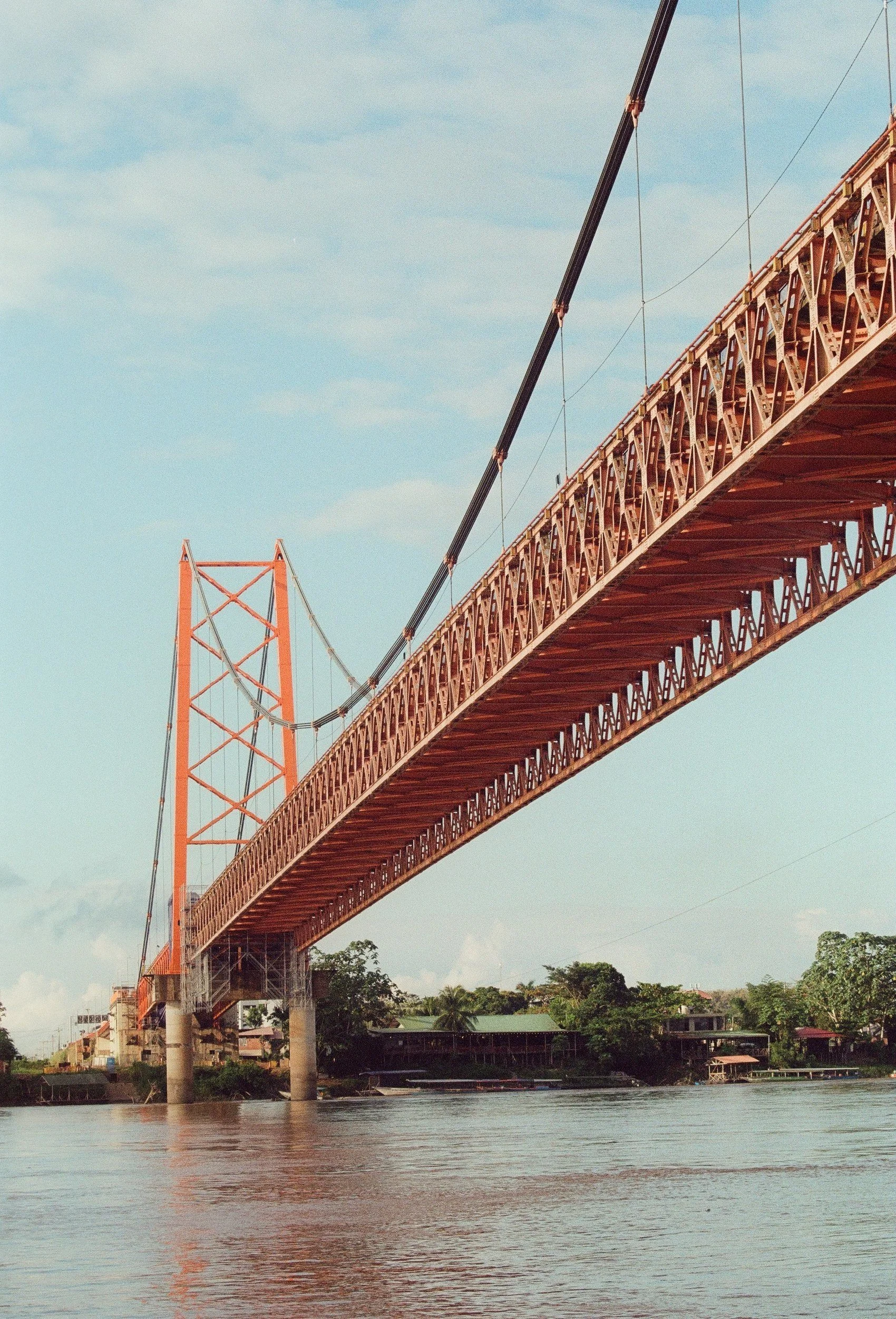 BRIDGE, the red suspension bridge over the river at Puerto Maldonado, Peru, in a film photograph by Omar Ali