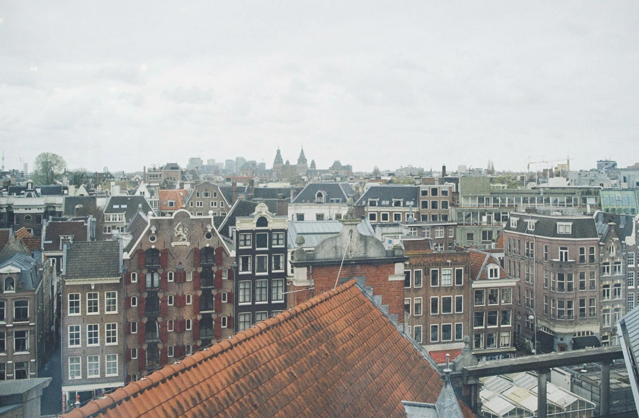 Cityscape with a mix of historic and modern buildings, including a prominent building with a white crest and red shutters, a red-tiled roof in the foreground, and cloudy sky.