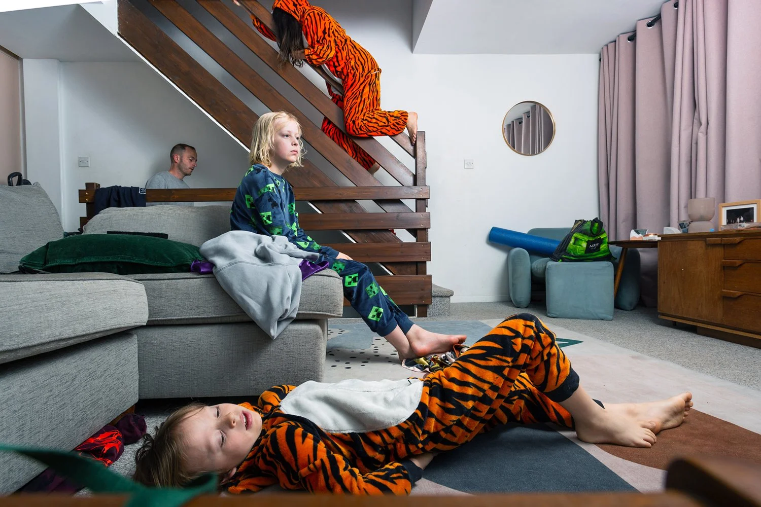 A family in pajamas, with one kid dressed in a tiger costume and a female in a tiger costume sliding on a living room near staircase bannister with a man in background, a young boy sitting on a sofa, and a boy lying on the floor on a rug.
