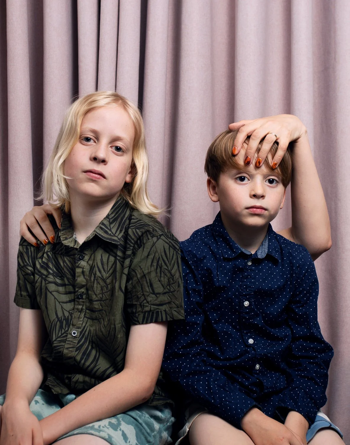 Two boys pose in smart shirt but casual shorts with adult hands with tiger pattern nails in front of a pale pink curtain