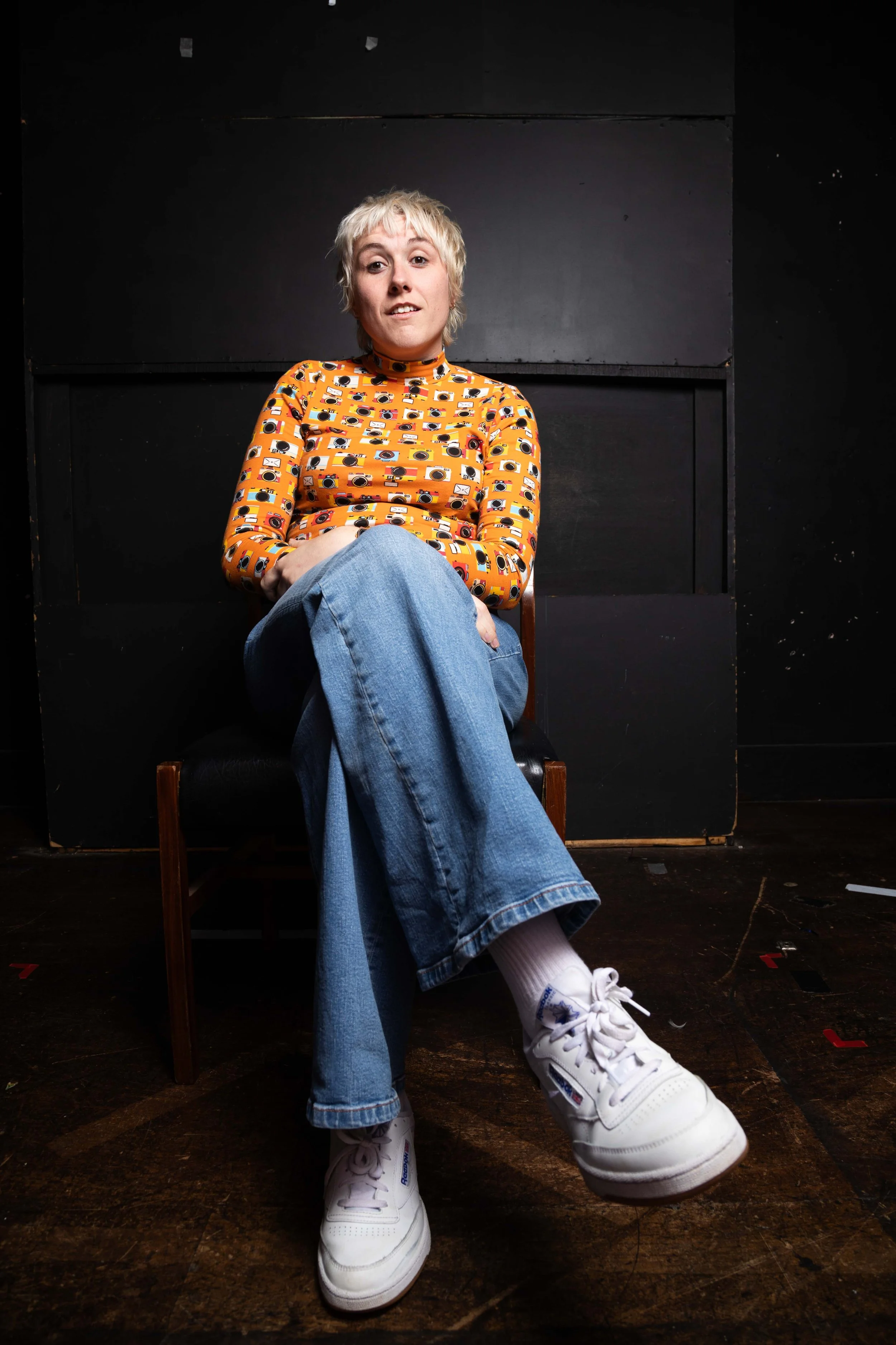 A female comedian wearing a mustard top with a camera pattern called Maisie Adam comedian photographed backstage in a black room by Karina Lax at The Comedy Village, Sheffield