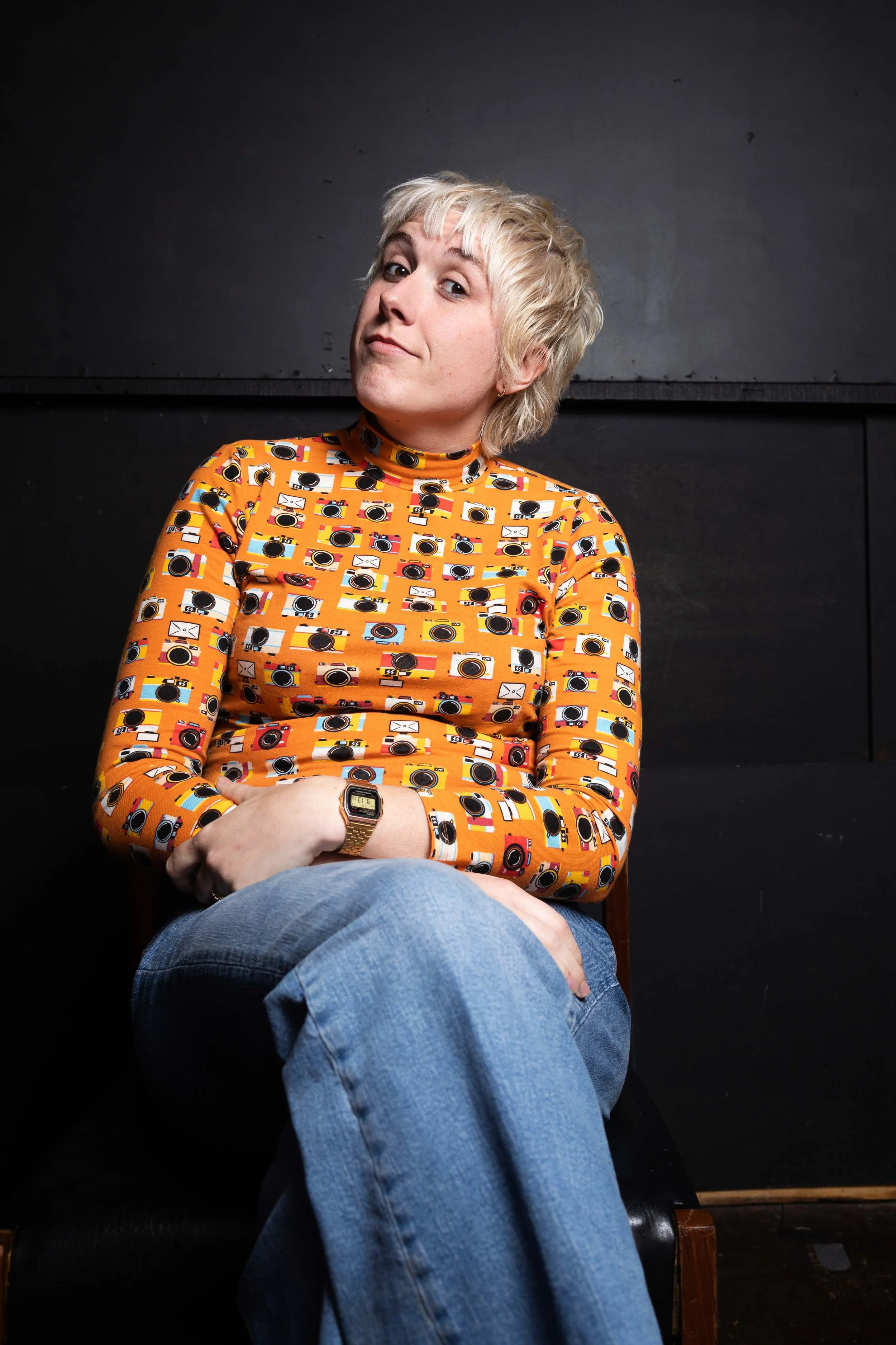 A female comedian wearing a mustard top with a camera pattern called Maisie Adam comedian photographed backstage in a black room by Karina Lax at The Comedy Village, Sheffield