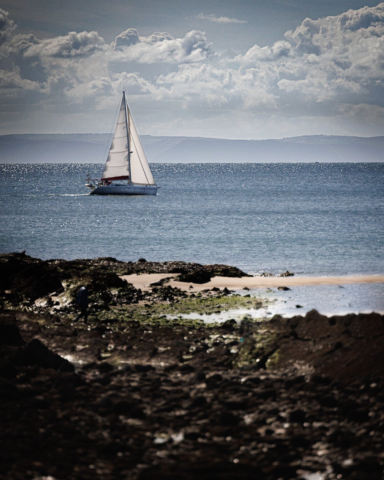 Sailboat on ocean with rocky shore in foreground and cloudy sky in background.