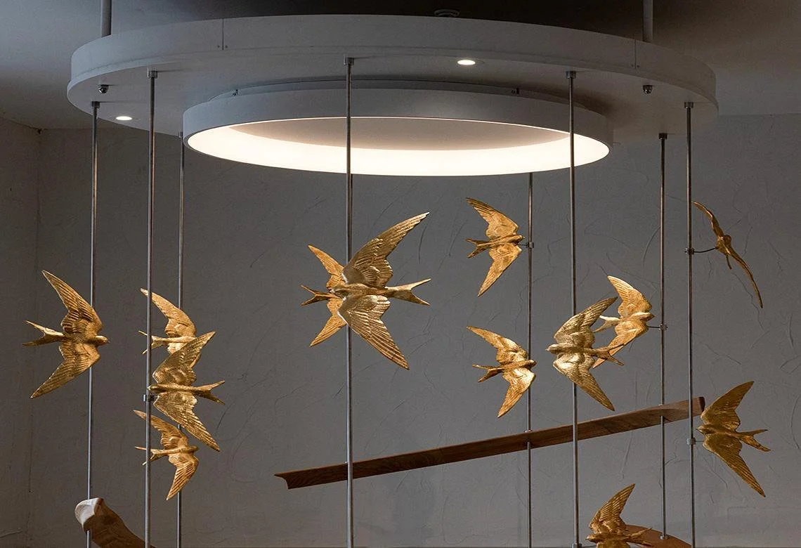 Gilded Swallow Sculpture Detail

A detailed view of a bespoke suspended sculpture, featuring gilded swallows arranged around a central light. The birds are mounted on polished metal rods, giving the piece a feeling of flight, movement and lightness w