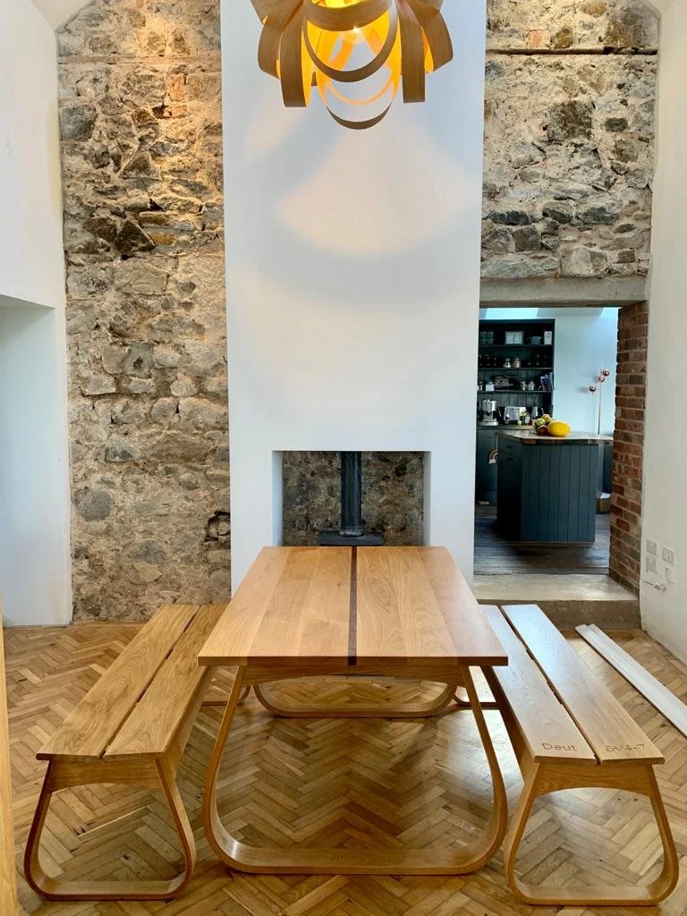 An oak table with steam bent legs and two matching benches in a room with exposed stone walls and a wood parquet floor. A modern hanging light fixture is above the table.