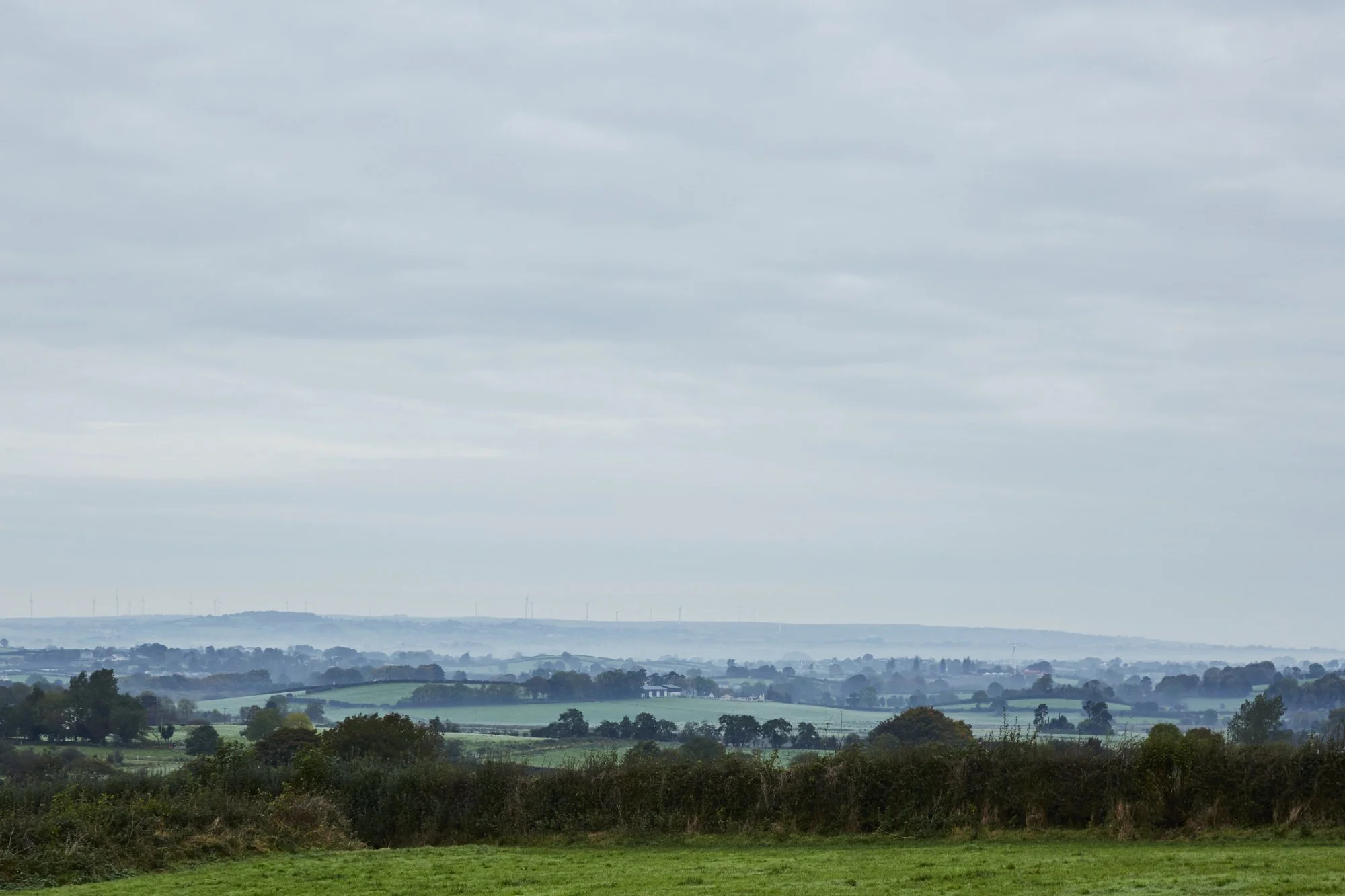 A wide view of a rolling countryside with green fields, scattered trees, and misty hills under a cloudy sky.