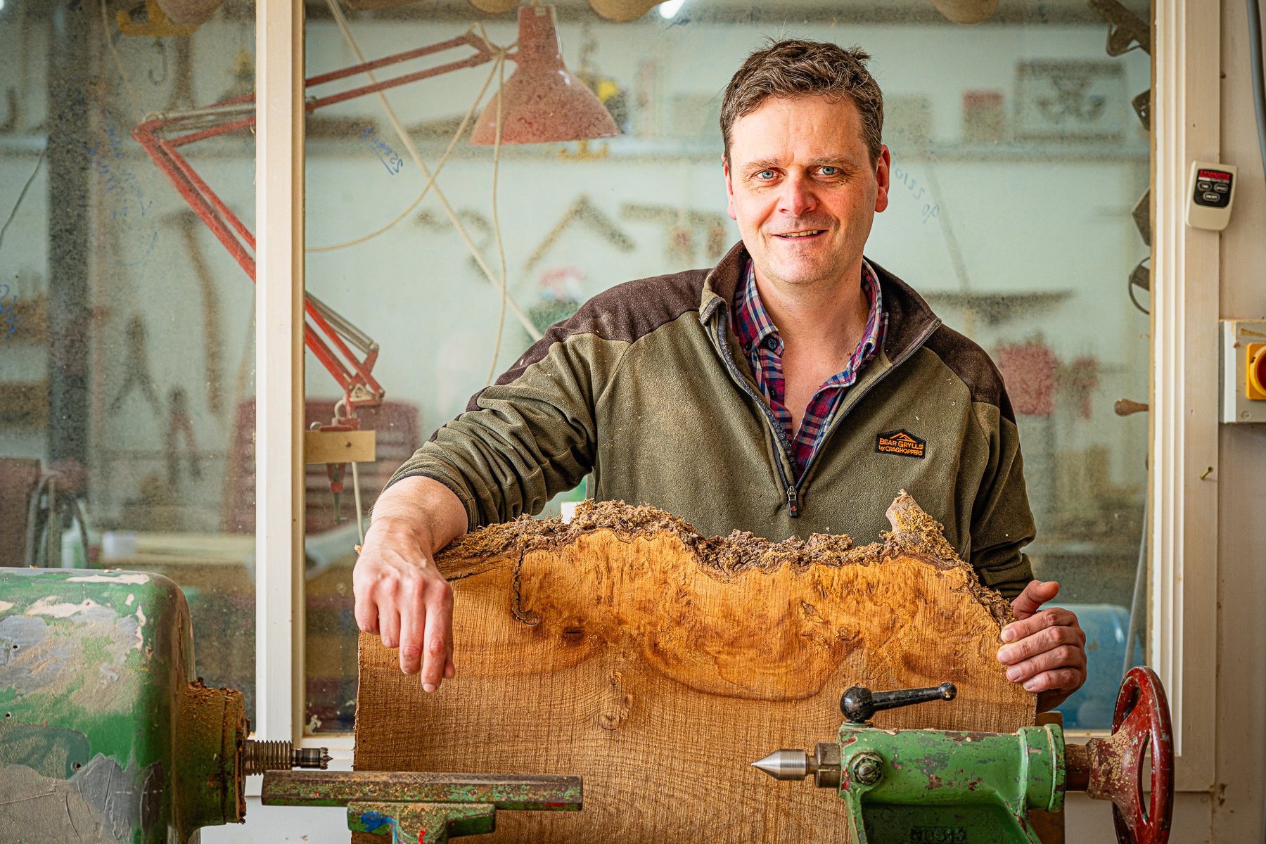 A man in a plaid shirt and brown jacket holding a large piece of wood in a woodworking shop.