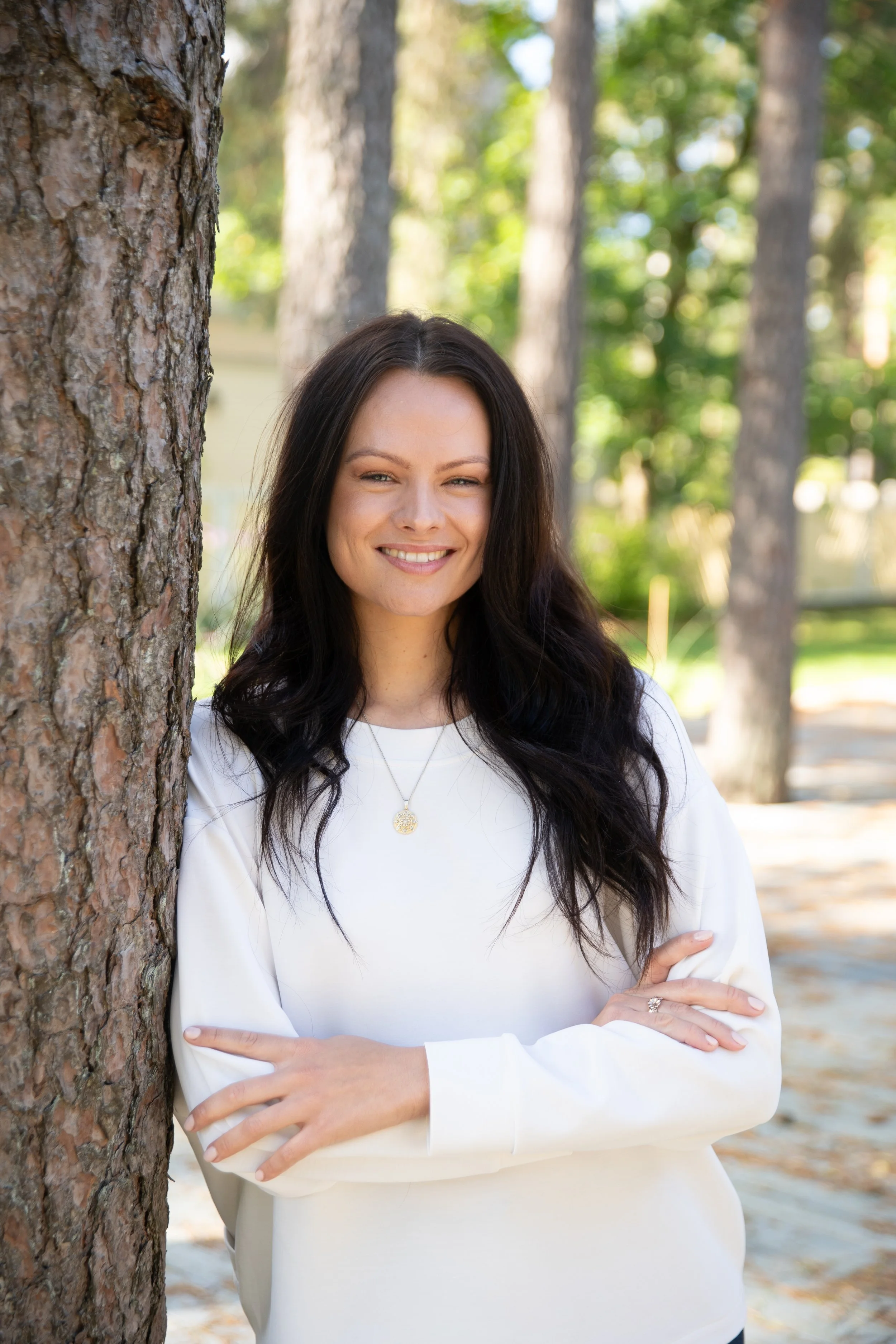 A woman with long black hair, smiling, leaning against a tree in a park with green foliage and trees in the background.