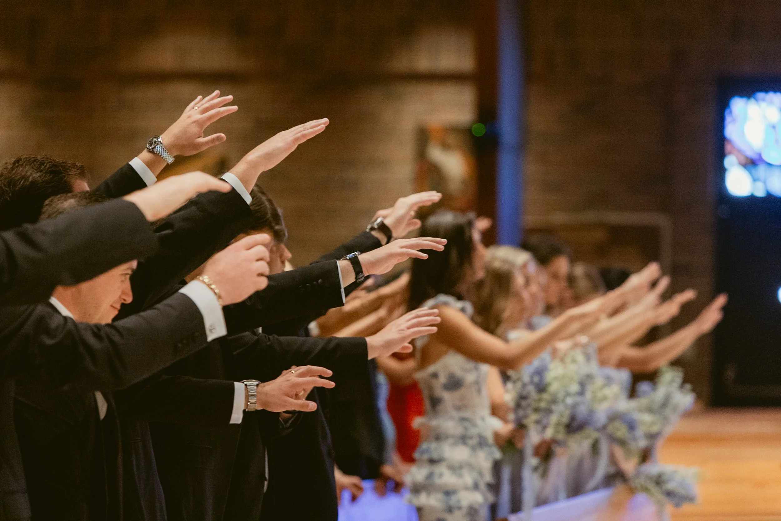 People at a wedding ceremony raising their hands during a prayer or blessing, with women holding bouquets in the background.