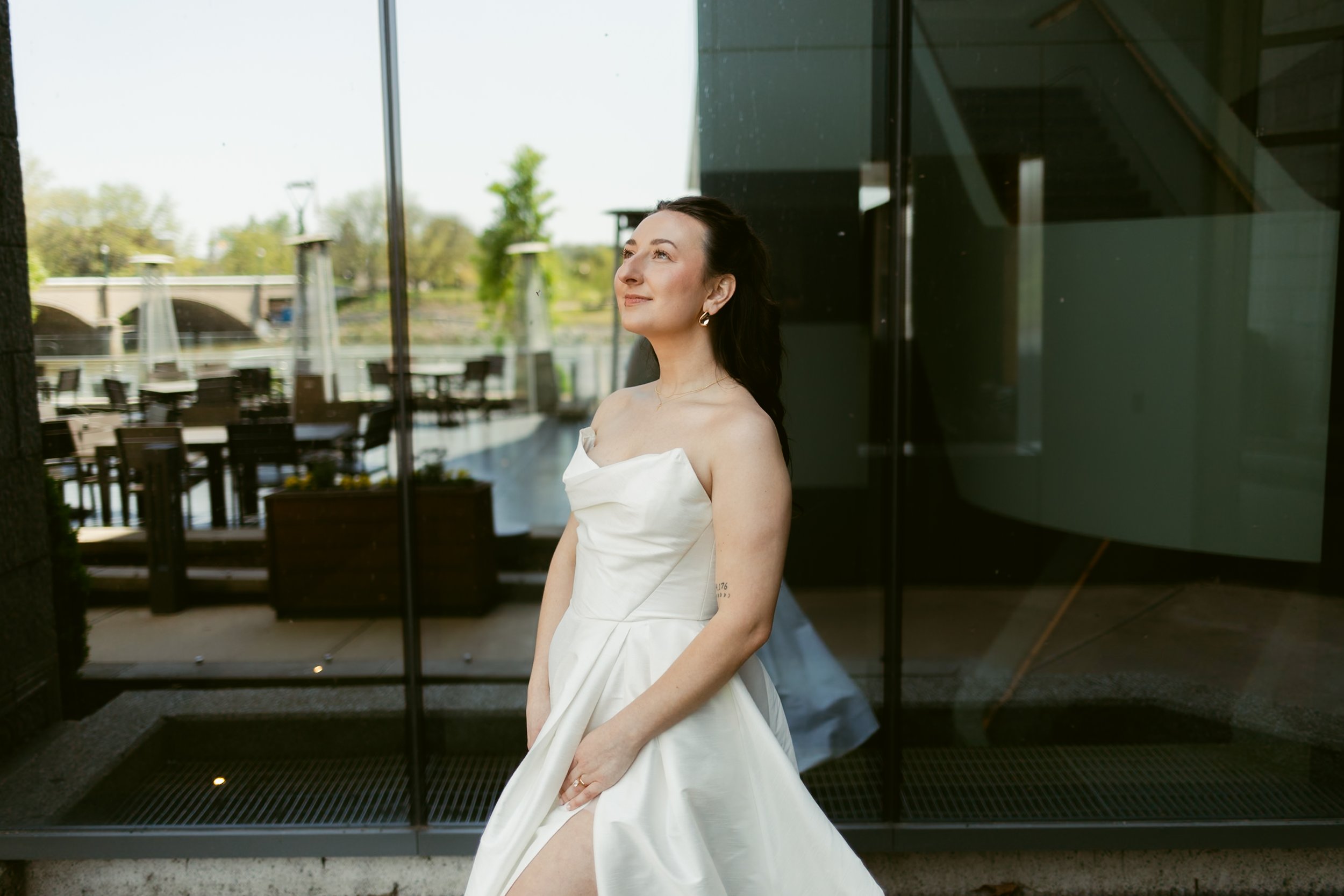Woman in a white wedding dress standing indoors near glass windows, with an outdoor patio and greenery visible outside.