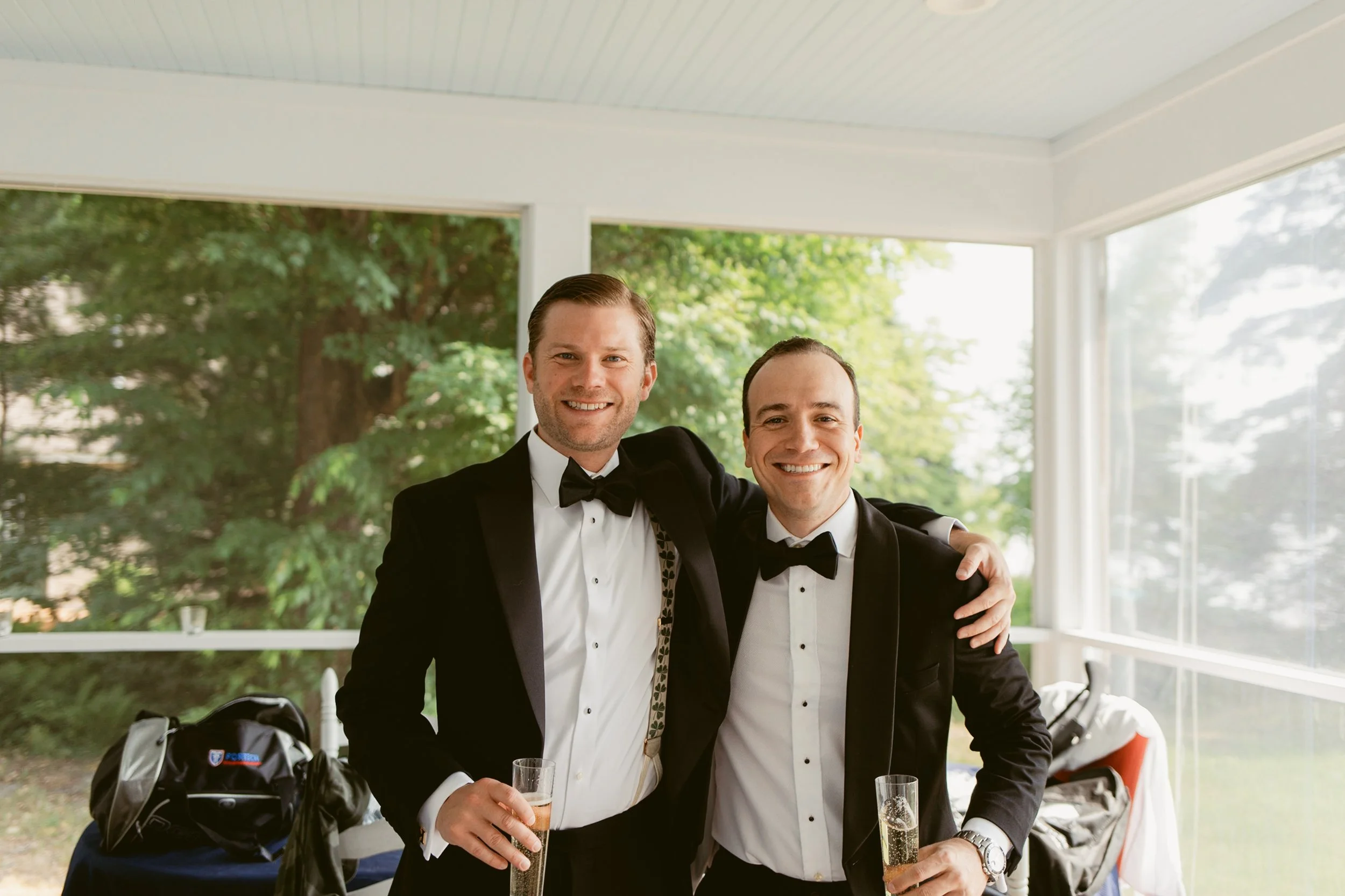 Two men in tuxedos smiling and holding champagne glasses, standing closely with one arm around each other, in a bright room with large windows and a view of green trees outside.