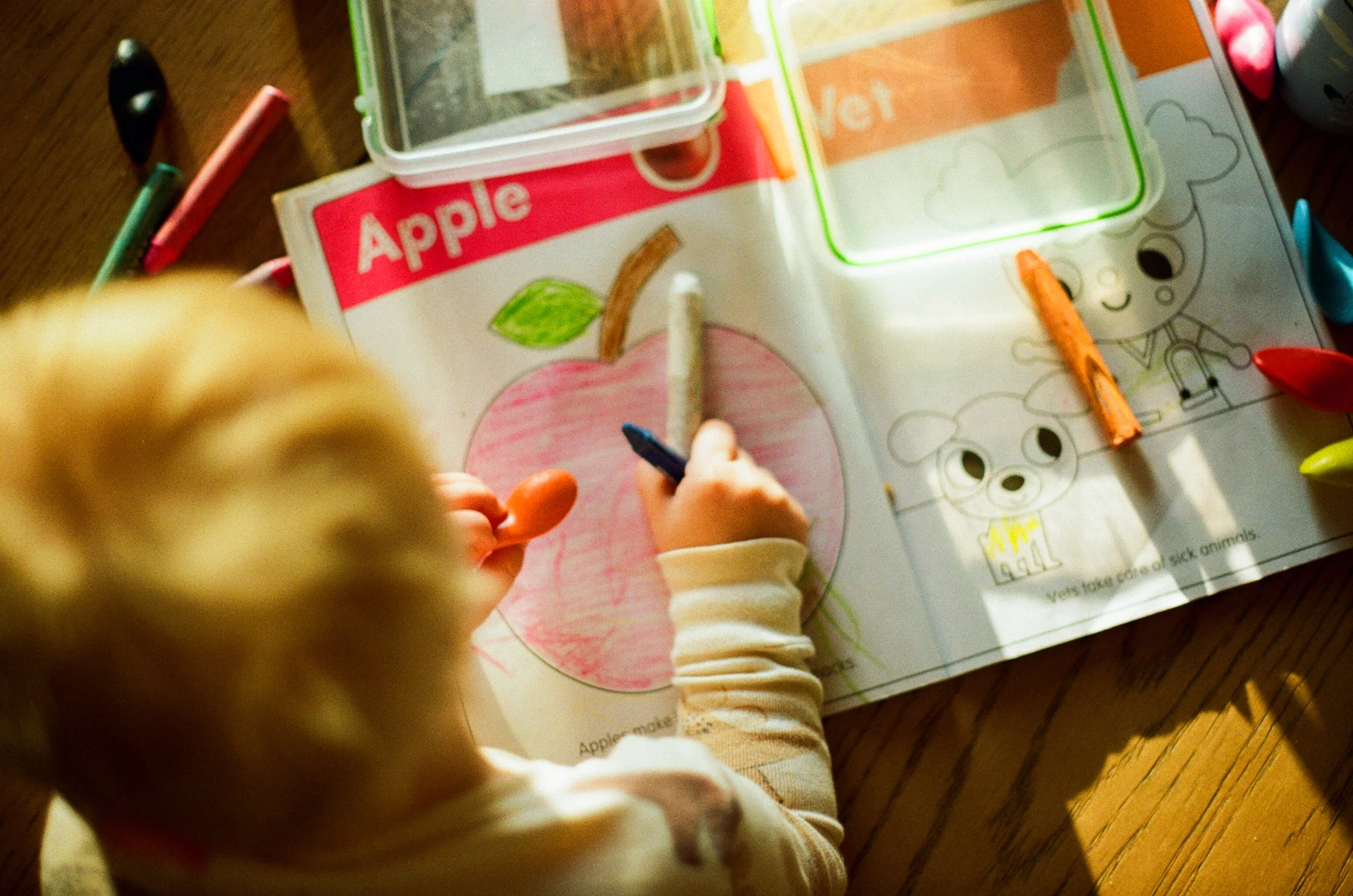 Child coloring a book with an apple illustration and various crayons on a wooden table.