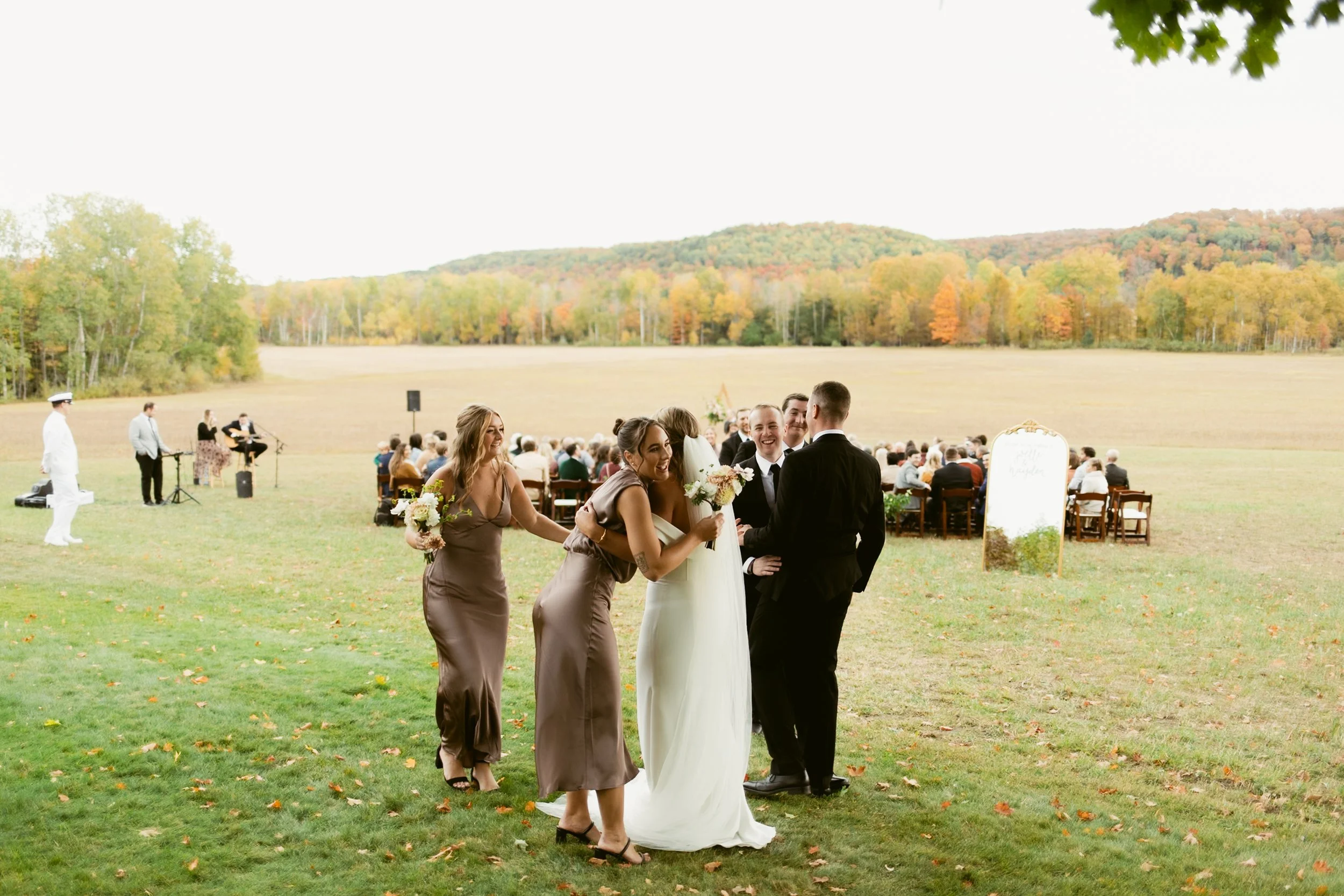 A wedding party standing on a grassy field with guests seated in the background, outdoors during fall with colorful trees on distant hills, featuring a bride, groom, two bridesmaids, and a small band with a singer.