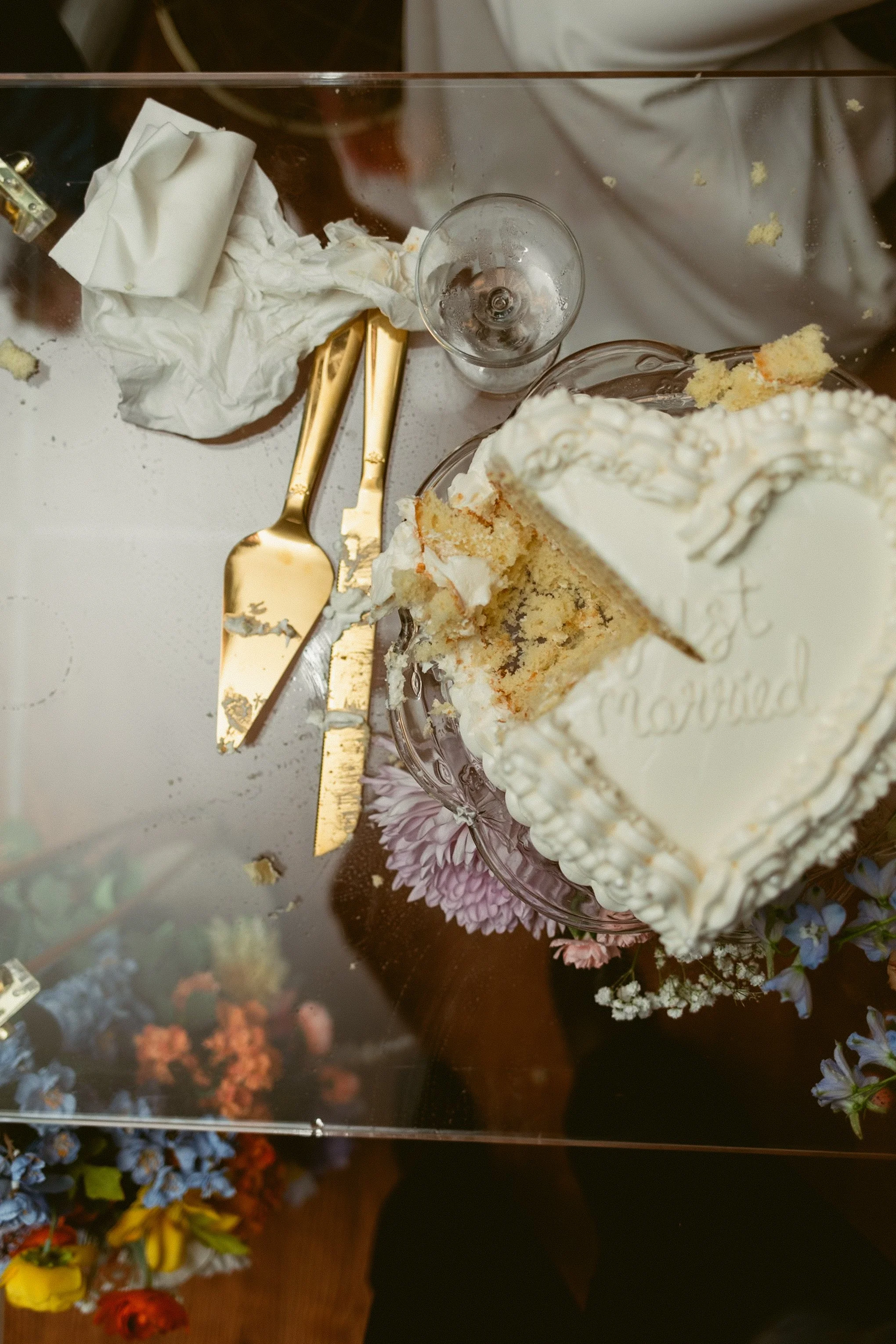 A crashed wedding cake with white frosting and floral decorations on a glass table, surrounded by gold cake serving utensils, an empty champagne glass, a crumpled napkin, and scattered cake crumbs. There are also flowers at the bottom edge of the tab