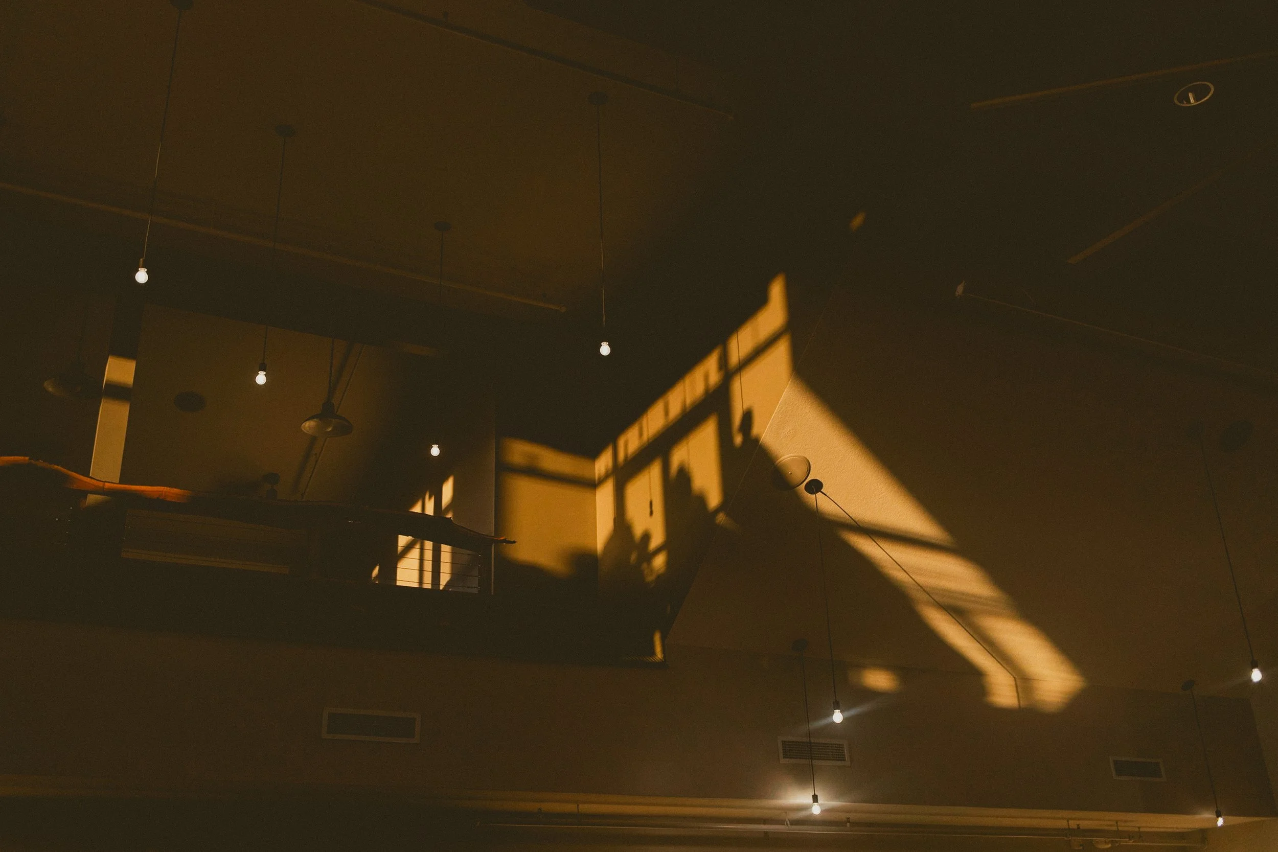 Sunlight casts shadows of a balcony railing and people onto a ceiling with hanging light bulbs in a dark room.