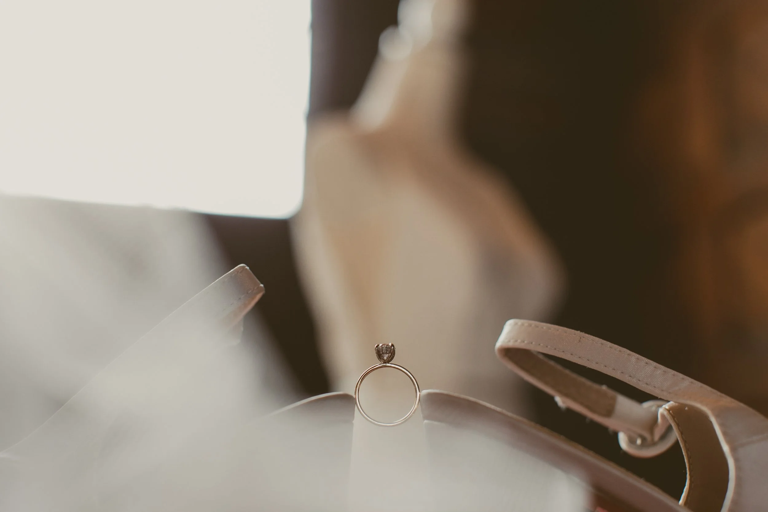 A close-up of a diamond engagement ring with a round stone, placed on a woman's finger, surrounded by beige and brown items, possibly shoes or accessories, with a blurred background and soft lighting.
