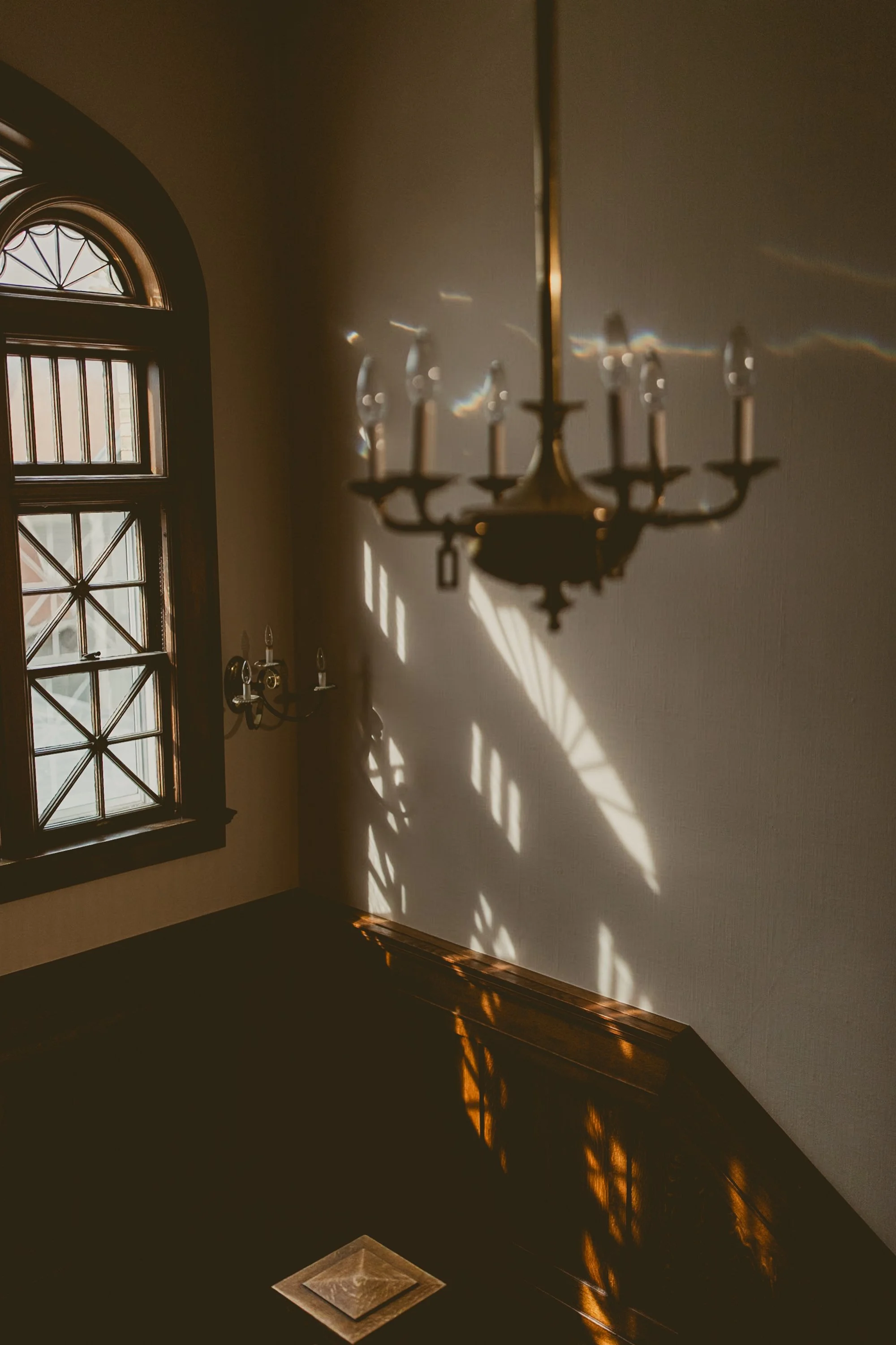 Sunlight streaming through a decorative window casting shadows on the wall, with a hanging chandelier visible overhead and a wall-mounted light fixture.