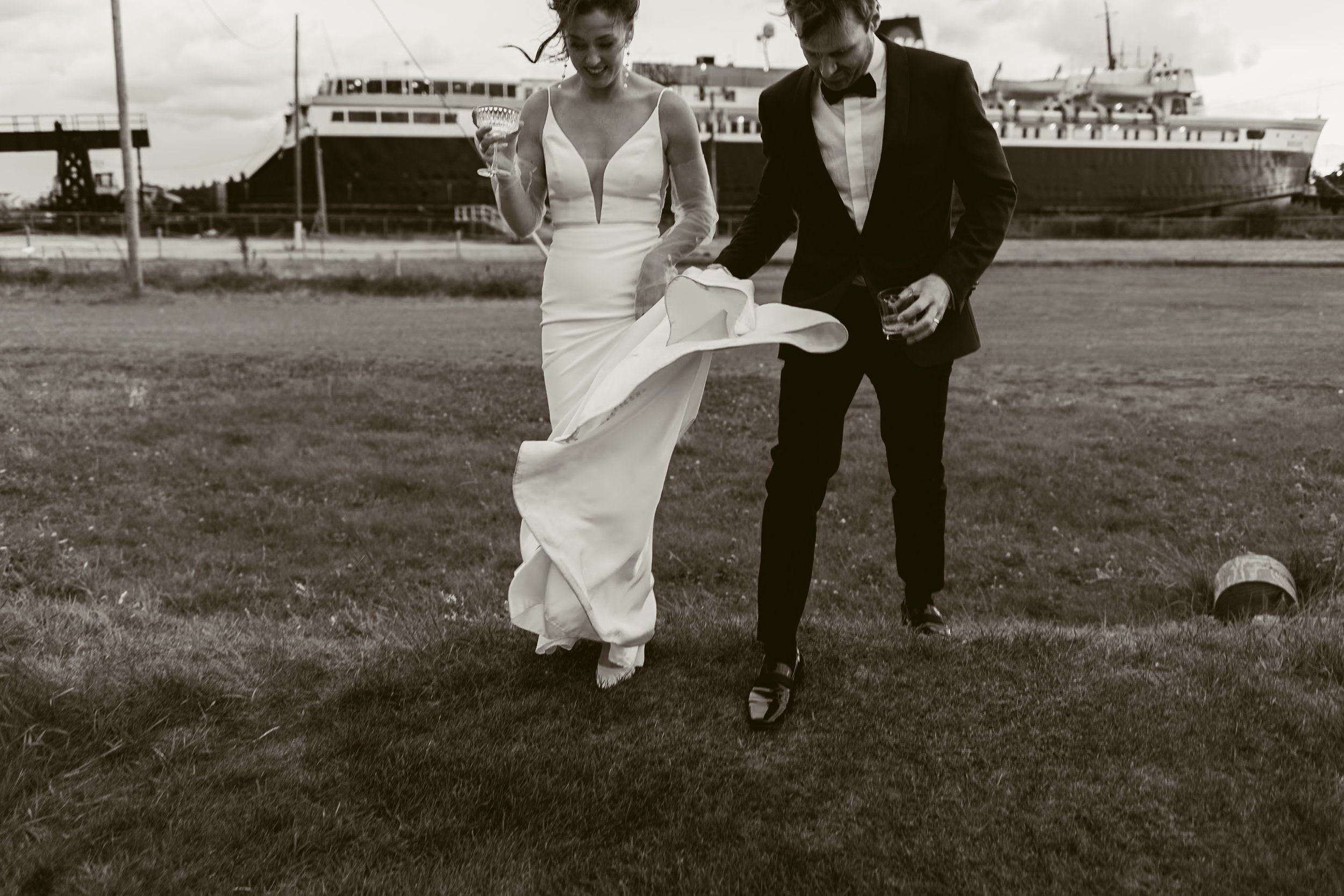 Couple in formal attire walking on grass, woman in a white wedding dress holding a glass, man in a tuxedo holding a drink, ship in the background.