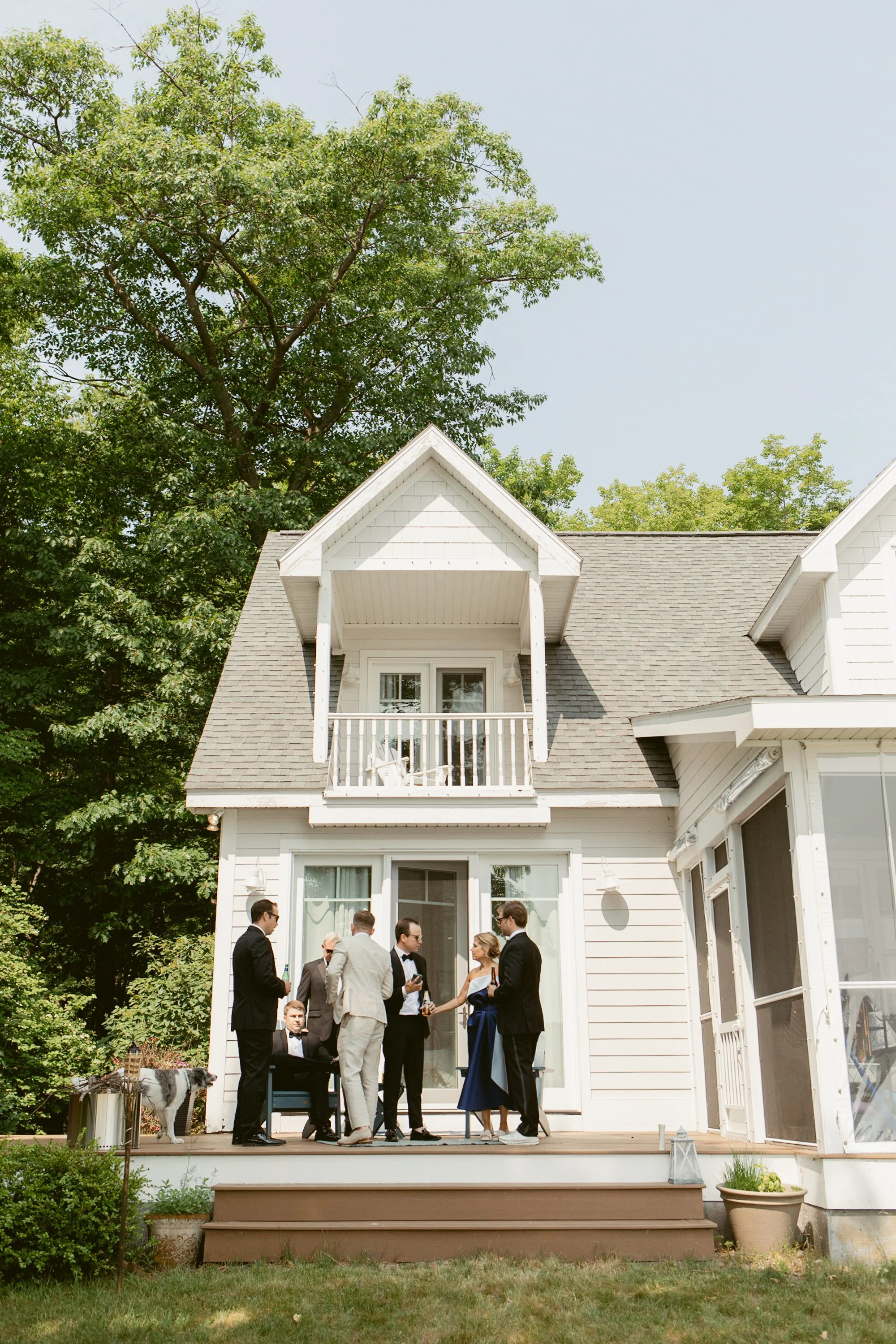 Group of people in formal attire gathered on a wooden deck outside a white house with a small balcony, surrounded by green trees during daytime.