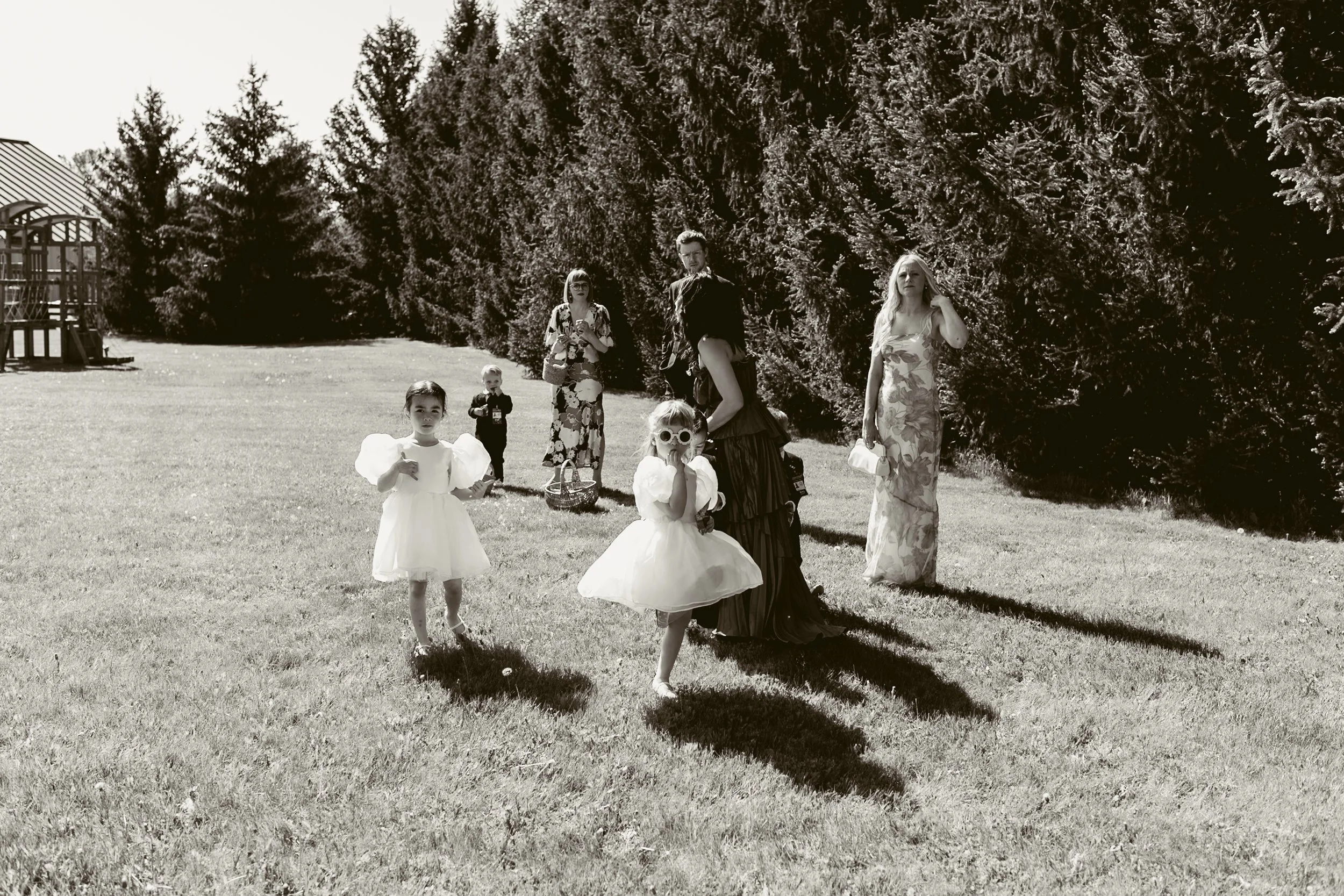 Children and women standing and walking on a grassy field with tall trees in the background, some dressed in formal or special occasion clothing, in black and white.