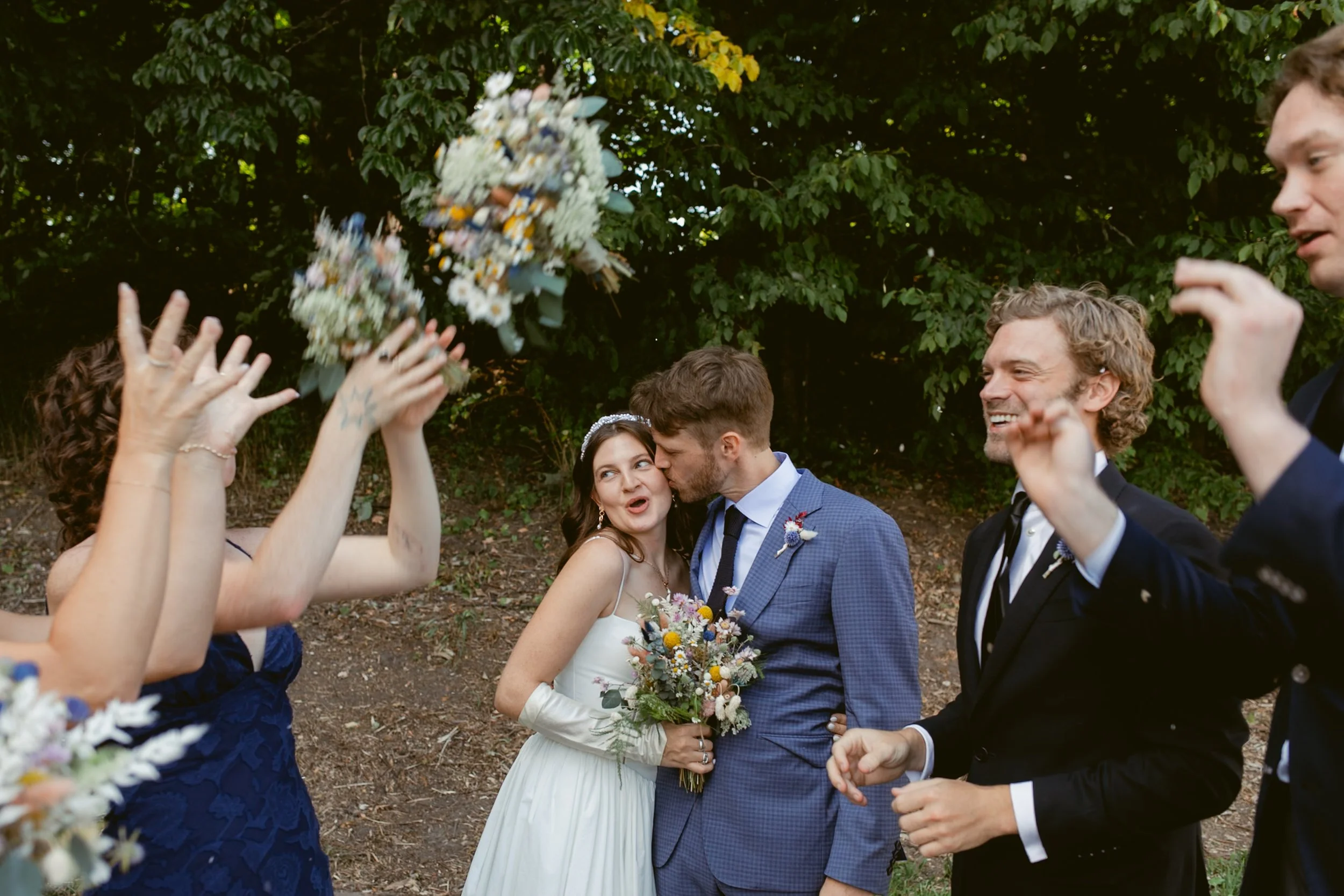A wedding scene with a bride and groom surrounded by friends in an outdoor setting. The bride is holding a bouquet and appears happy while the groom is kissing her on the cheek. Friends are celebrating, with some throwing flower bouquets in the air.