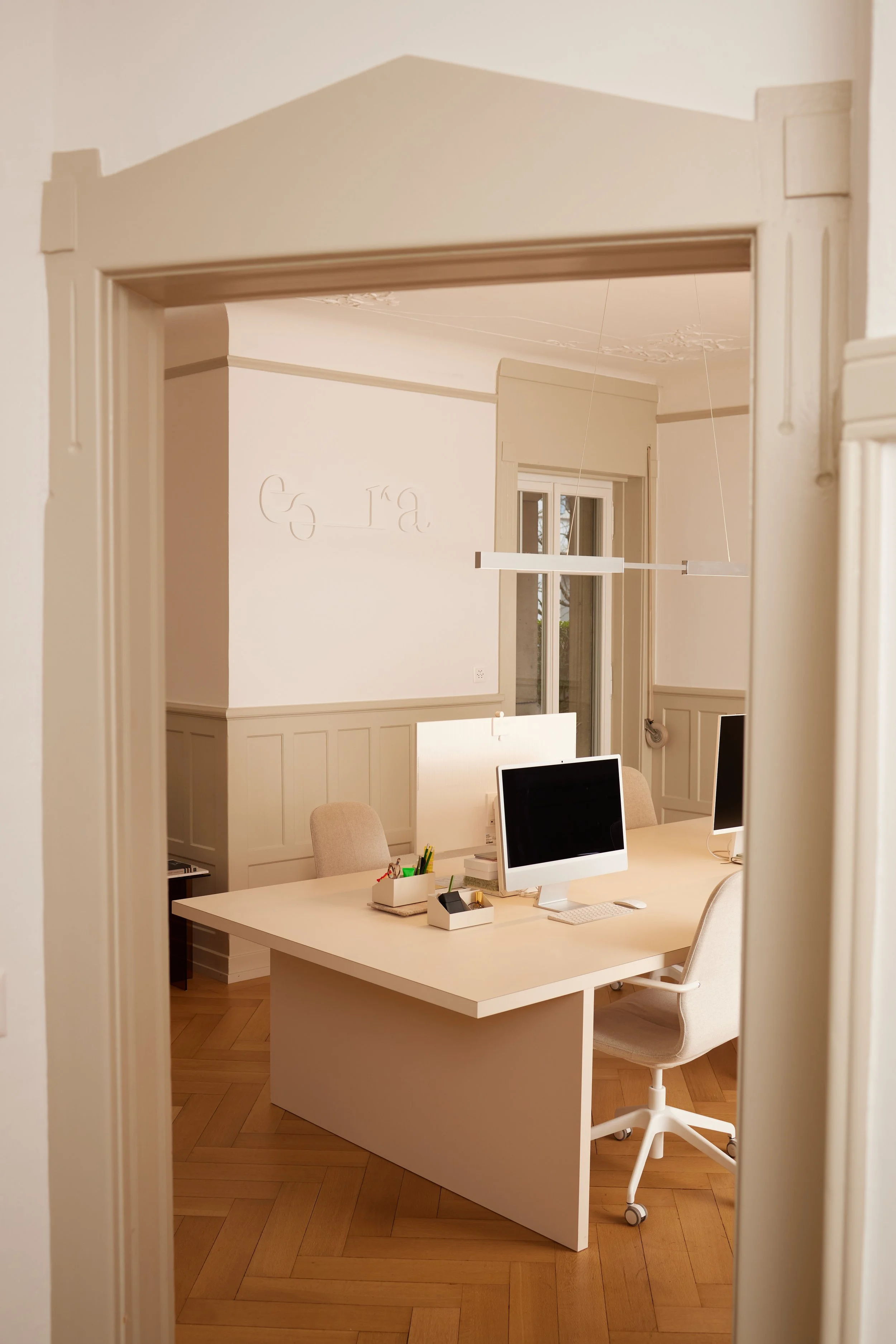 Ein modernes Büro mit einem großen weißen Schreibtisch, auf dem zwei Computerbildschirme, Schreibutensilien und ein kleiner Organizer stehen. Es gibt einen ergonomischen weißen Bürostuhl und im Hintergrund große Fenster, die viel Tageslicht ins Zimmer lassen. Die Wände sind hell gestrichen mit dekorativen Elementen und Wandschriften.