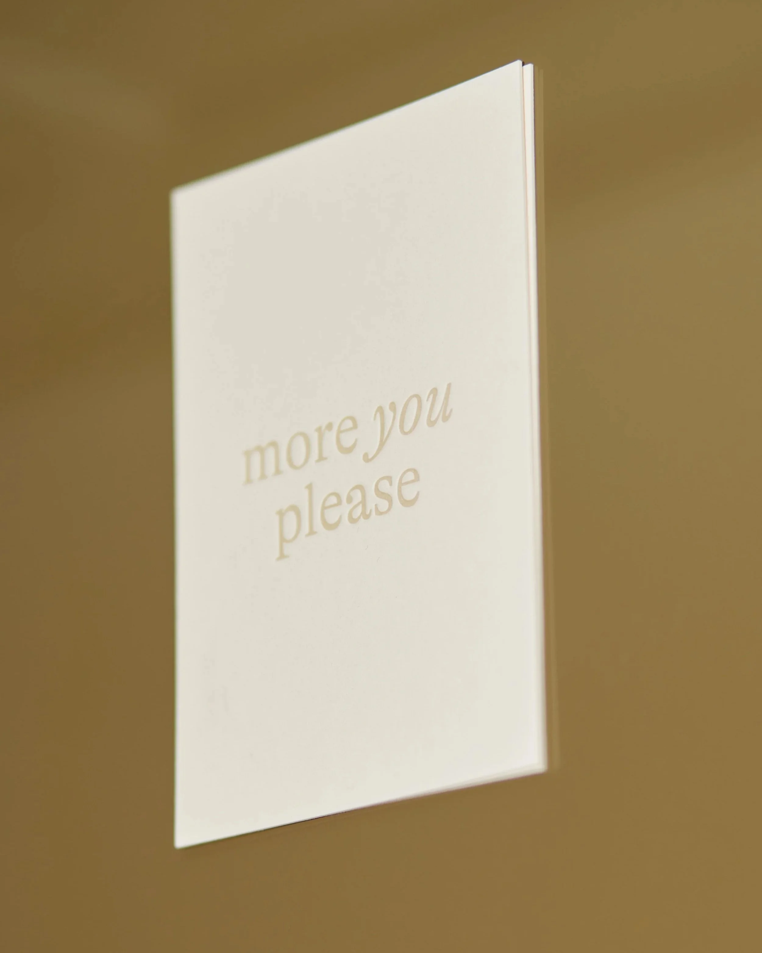 Hellerweise Karte mit Text 'more you please' auf weißem Hintergrund, auf braunem Tisch liegend.