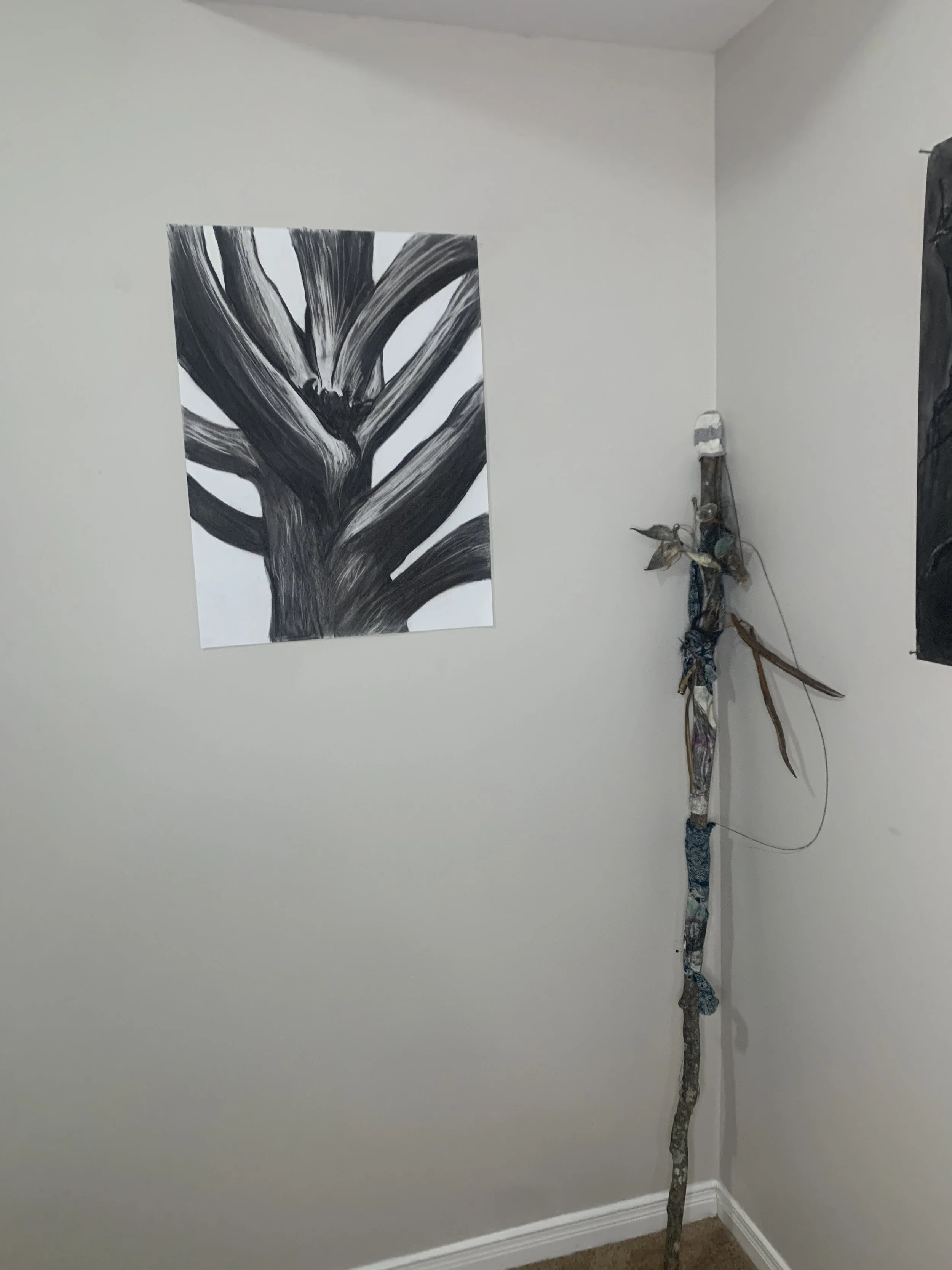 A corner of a room with a large black and white painting of a tree on the wall. In the corner, there is a tall, thin, natural-looking branch leaning against the wall, with some fabric and wire decorations attached to it.