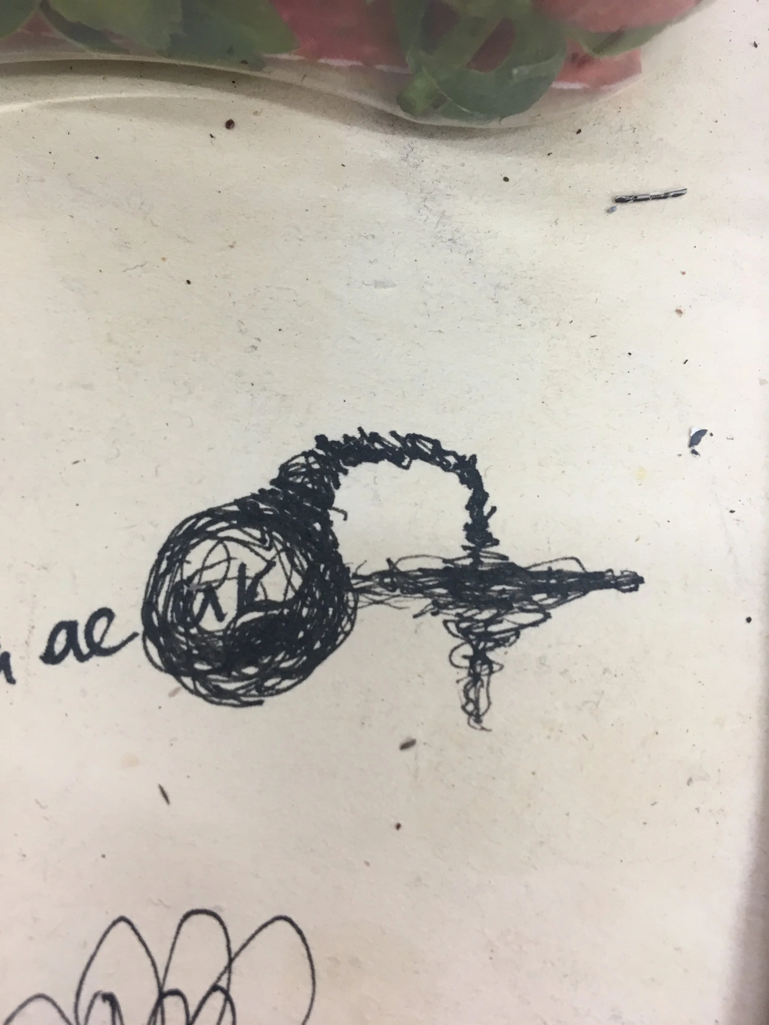 Close-up of a handwritten doodle of a black swirly bird or animal on a white piece of paper with some scattered dirt and ink smudges on the surface and a colorful object partially visible at the top.