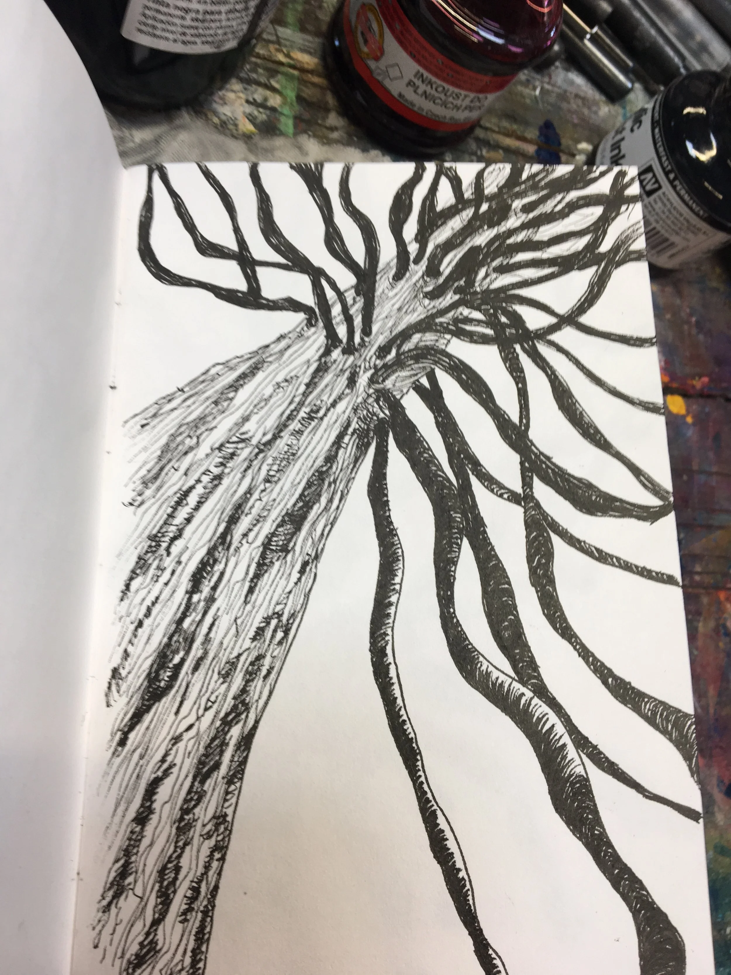 Black ink drawing of a tree with numerous twisting and curving branches on white paper, surrounded by art supplies and bottles on a work surface.