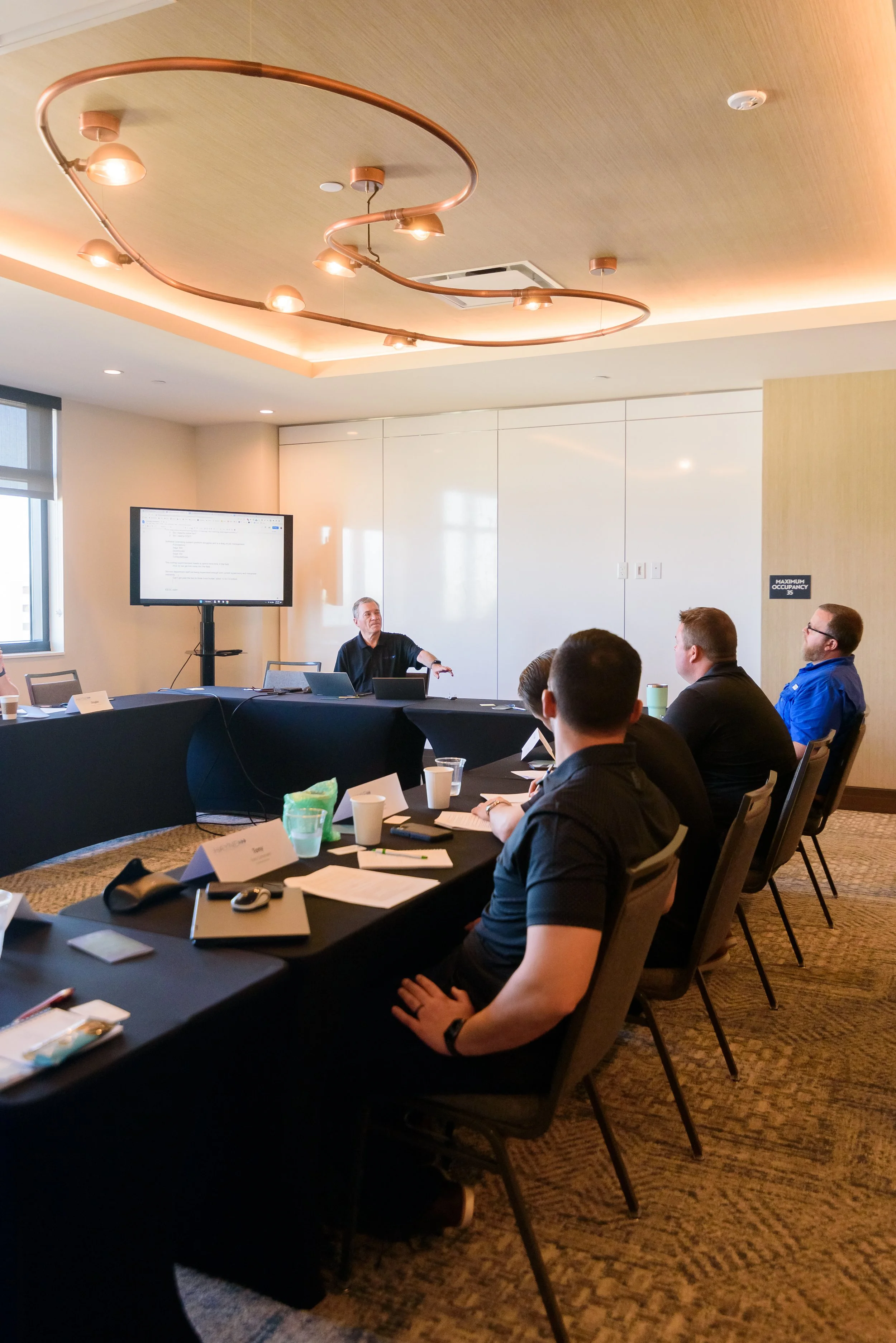Business meeting with multiple people seated around a conference table, one person presenting with a screen displaying information.