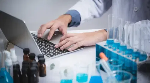 Person working on a laptop in a laboratory with test tubes and medication bottles on the desk.