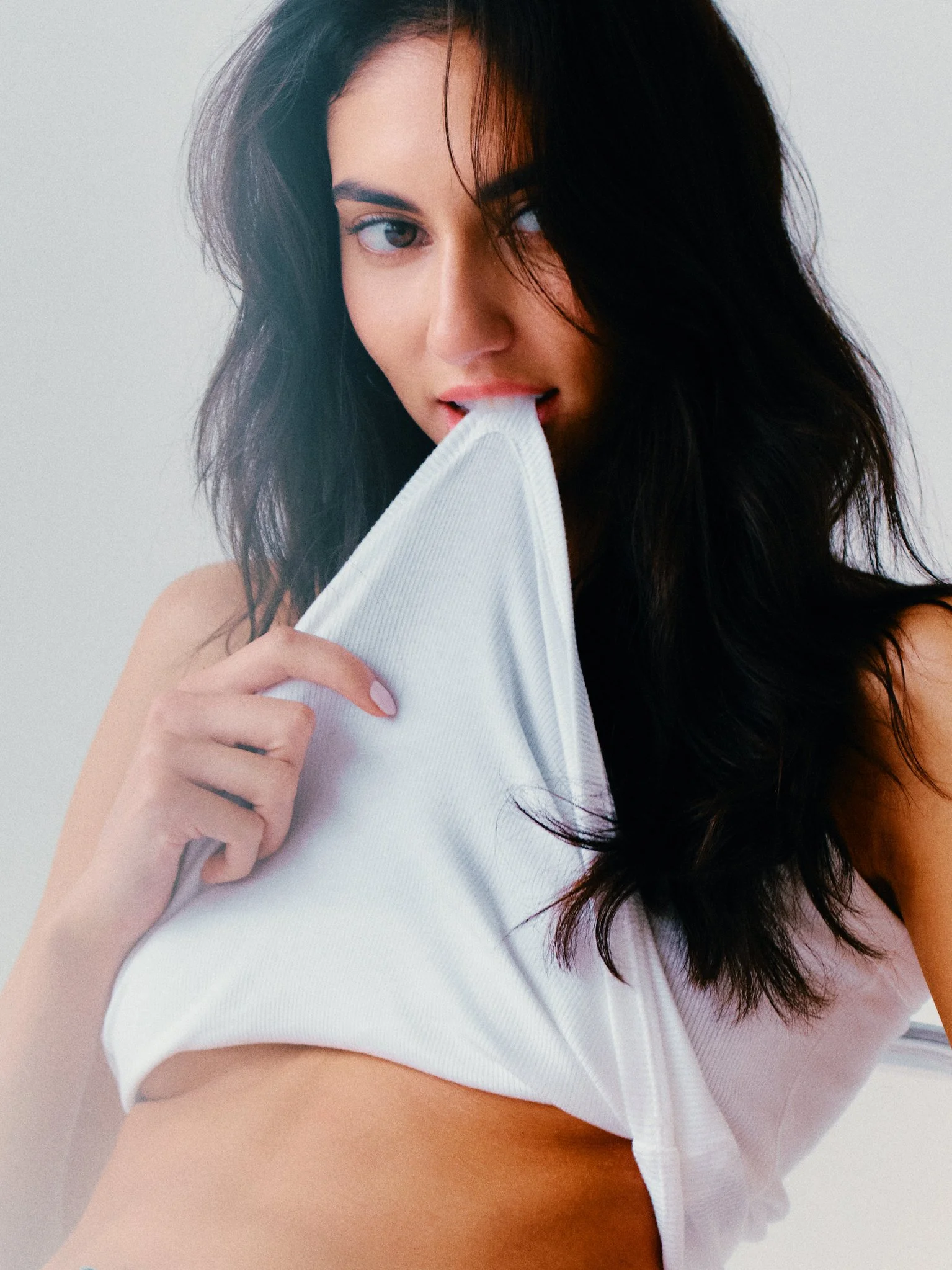 A woman with dark, wavy hair holding a white shirt near her mouth while looking at the camera.