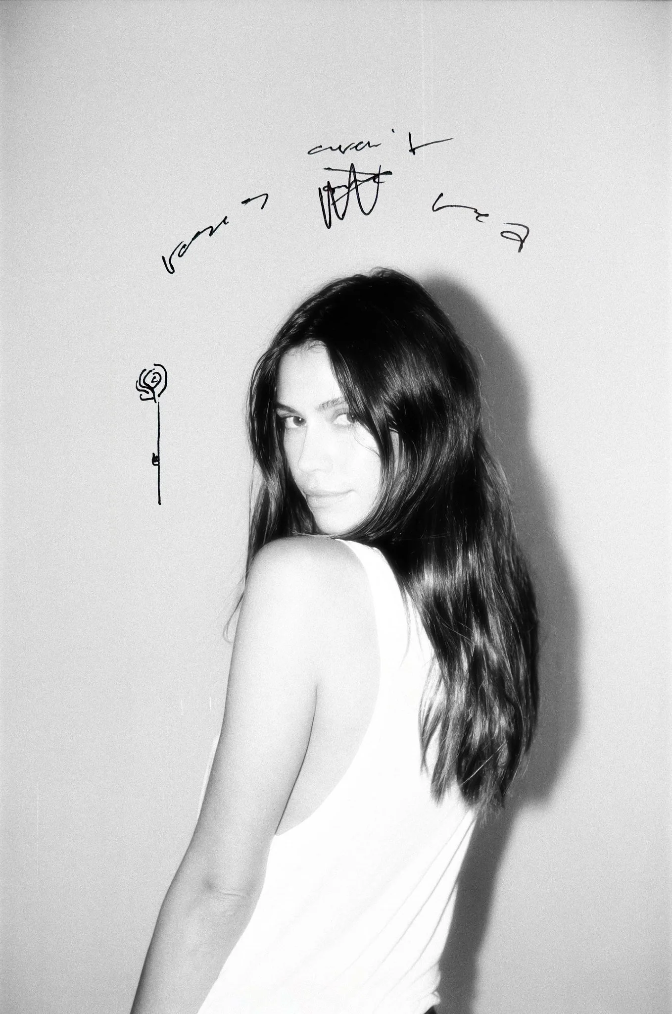 Black-and-white photo of a woman with long dark hair, partially turned, with handwritten text and a small drawing of a flower on the wall behind her.