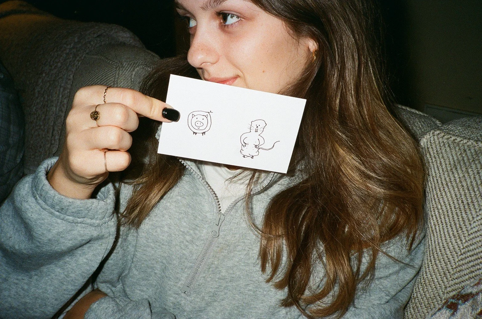 A woman with long brown hair, wearing a gray zip-up hoodie, is holding a small white card with cartoon drawings of a pig and a mouse near her face, partially covering her mouth.