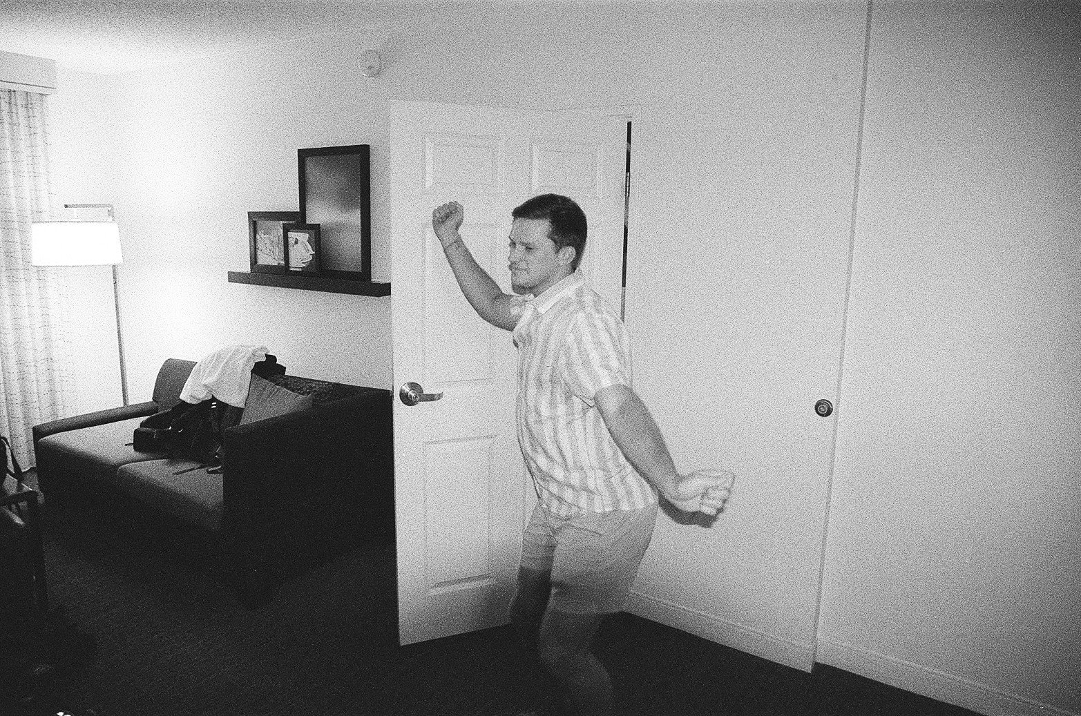 A man dancing in a hotel room with a couch, a standing lamp, and a window with curtains.