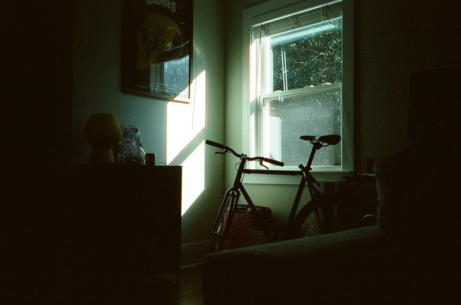 A dimly lit room with sunlight coming through a window, casting shadows on the wall. There is a bicycle leaning against the wall near the window, and a dresser or table with a lamp, a few objects, and framed pictures on it.