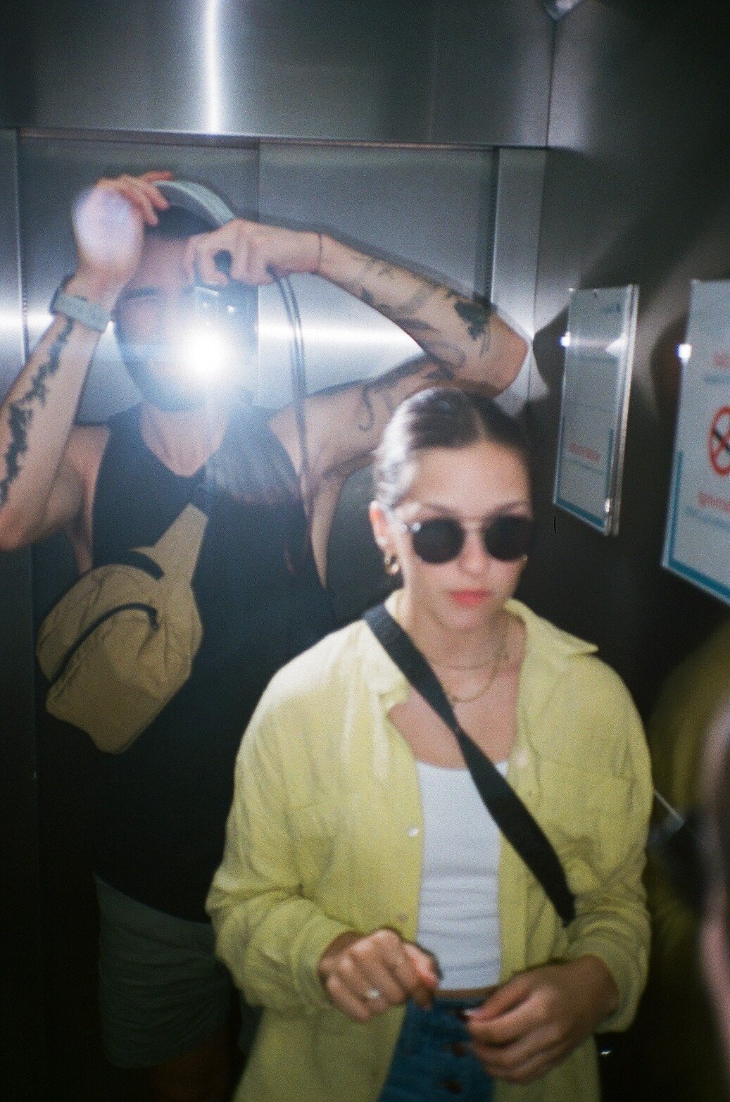 Two women in an elevator taking a selfie, one wearing sunglasses and a yellow jacket, and the other on the mirror with tattoos taking the photo.