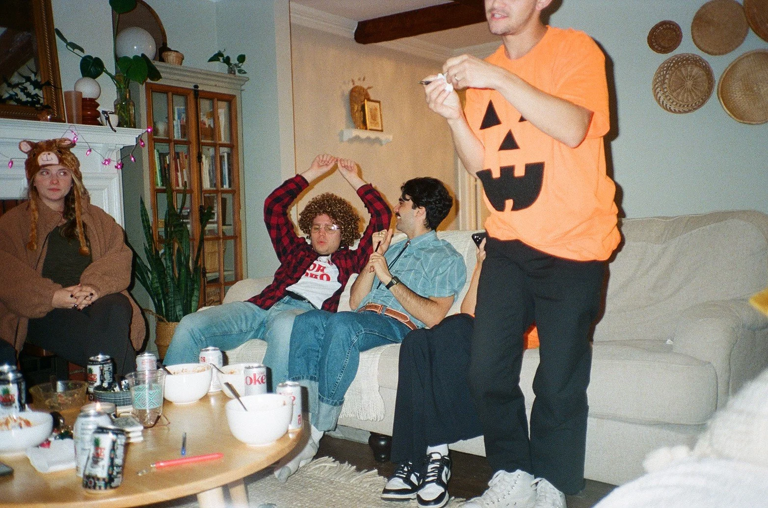 Group of people at a Halloween party, with some dressed in costumes like a pumpkin shirt and a bear hat, sitting on a cream-colored couch and socializing in a cozy living room.