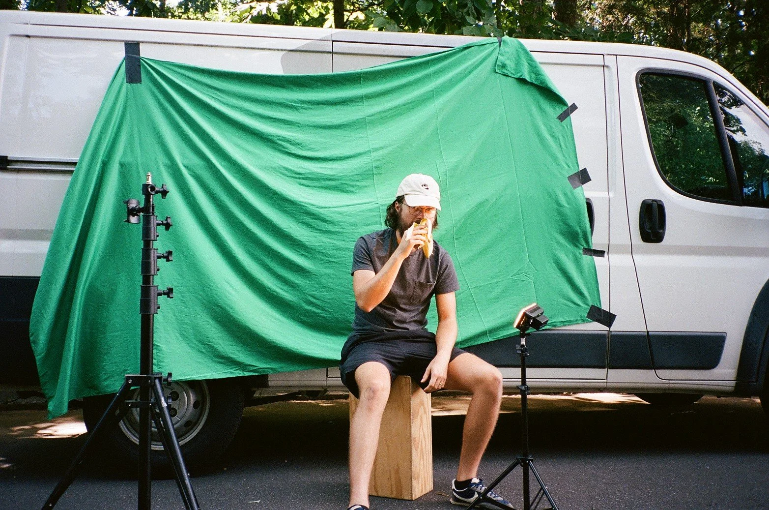 Person sitting on a wooden block in front of a white van with a green screen backdrop, eating a sandwich, with lighting equipment around him.