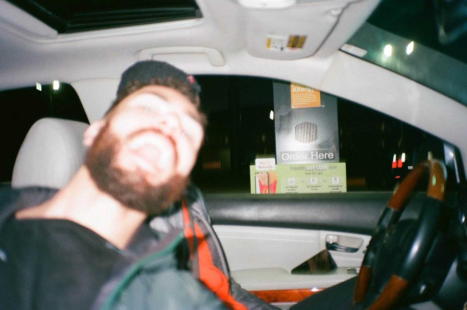 A man with glasses and a beard smiling or laughing in the driver's seat of a vehicle at night, with a fast-food order sign visible outside the window.