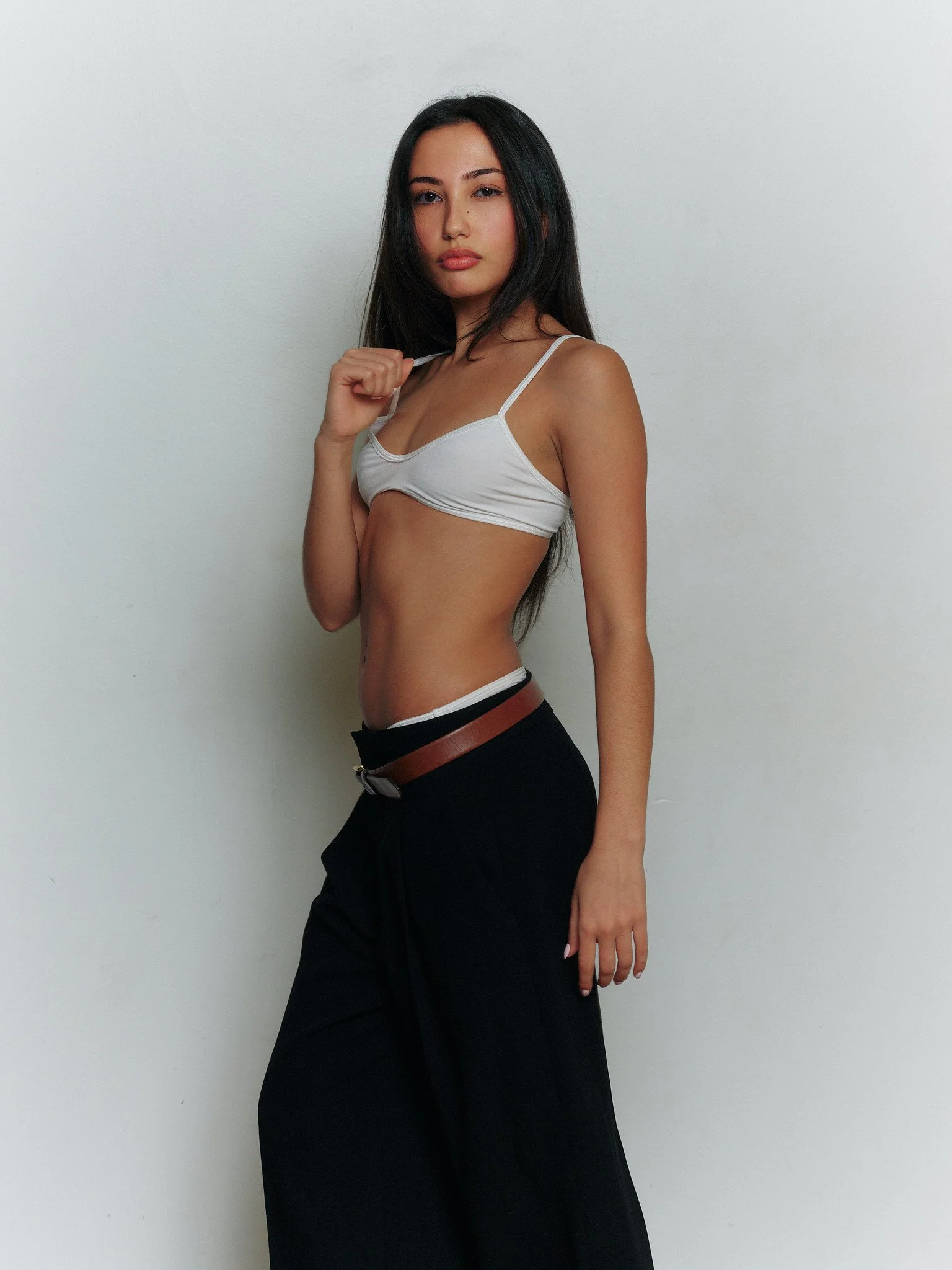 Young woman with long dark hair wearing a white crop top, black wide-leg pants, and a brown belt, posing against a plain white wall.