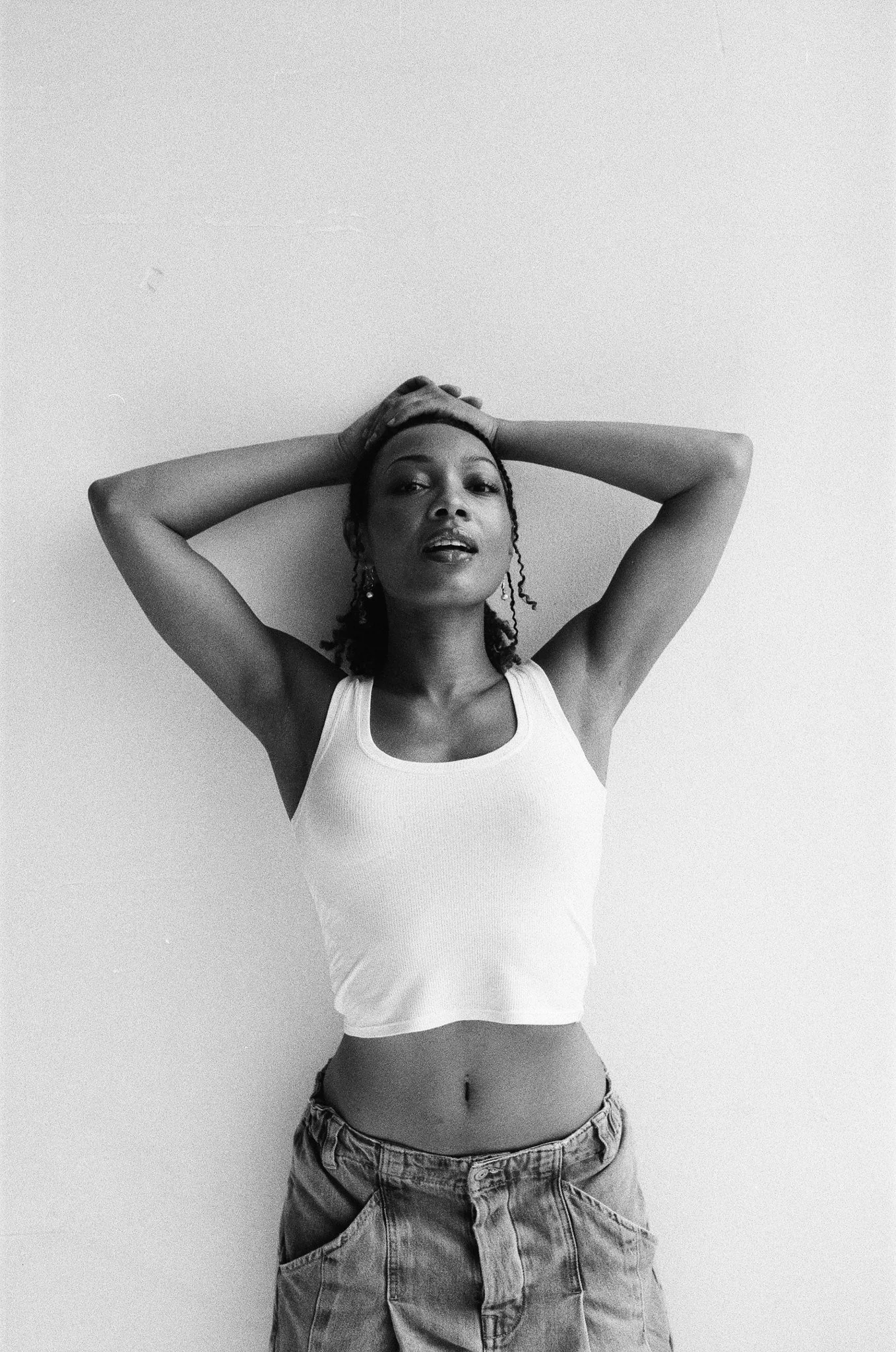 Black and white photo of a woman wearing a white tank top and baggy denim pants, posing with hands on her head against a plain wall.