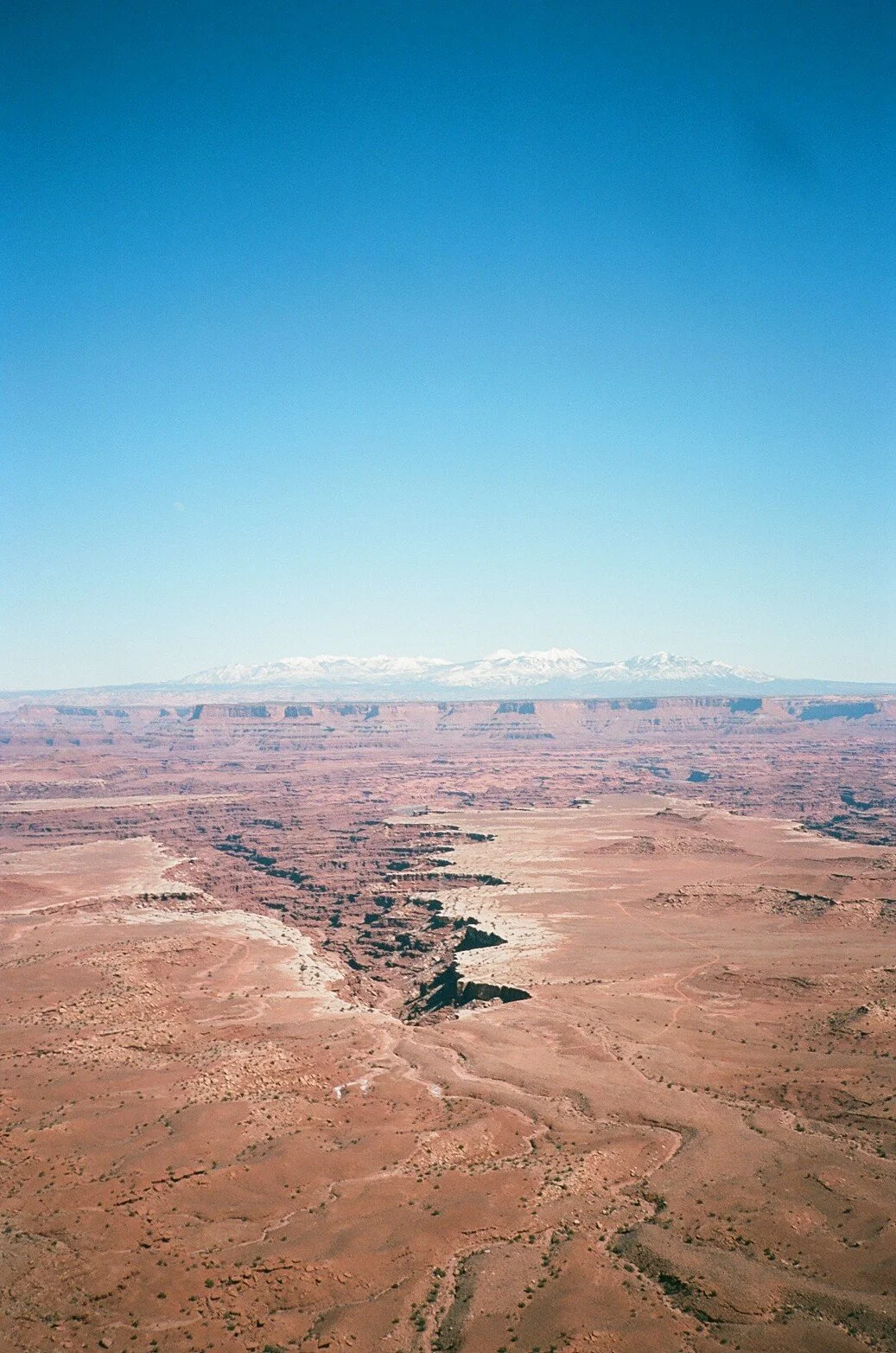 Aerial view of a desert landscape with a deep canyon, distant snow-capped mountains under a clear blue sky.