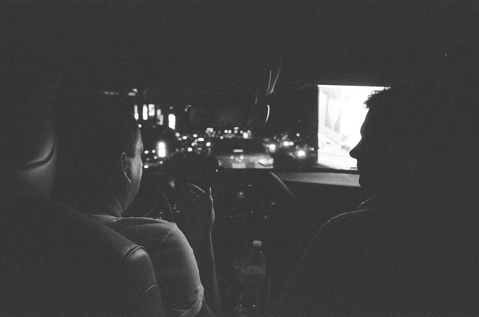 Two people sitting in the front seats of a car at night. The driver is looking at the road, and the passenger is facing forward, with city lights visible through the windshield.