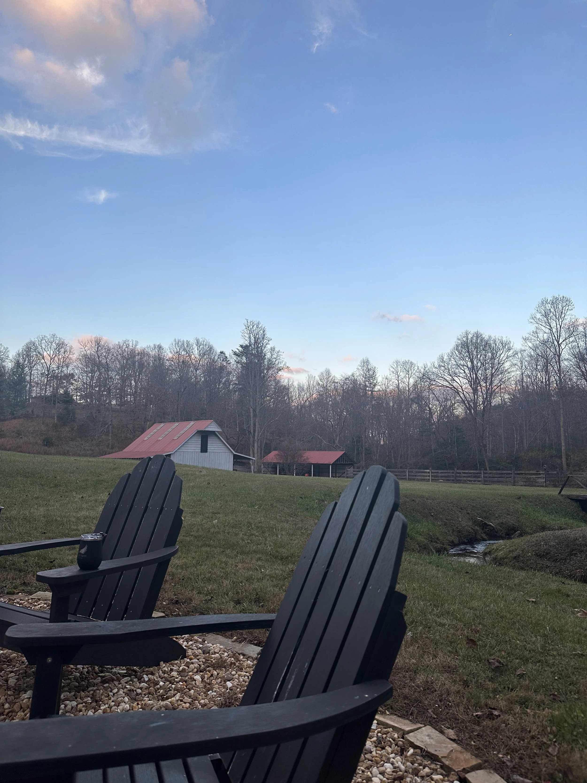 Two black Adirondack chairs on a gravel patch in a grassy yard, overlooking a small creek, with a rustic barn and trees in the background, under a clear blue sky.